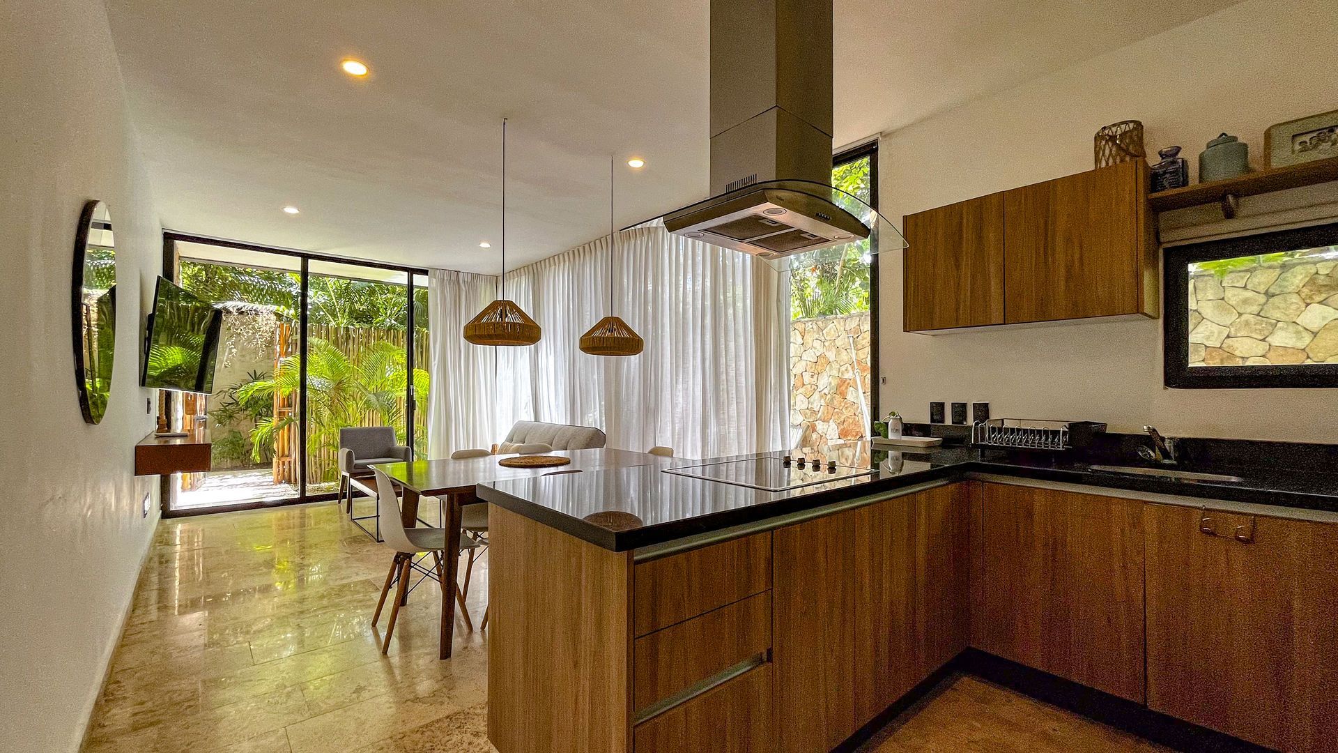 Cocina moderna con gabinetes de madera, isla y grandes puertas de vidrio que dan a la vegetación.