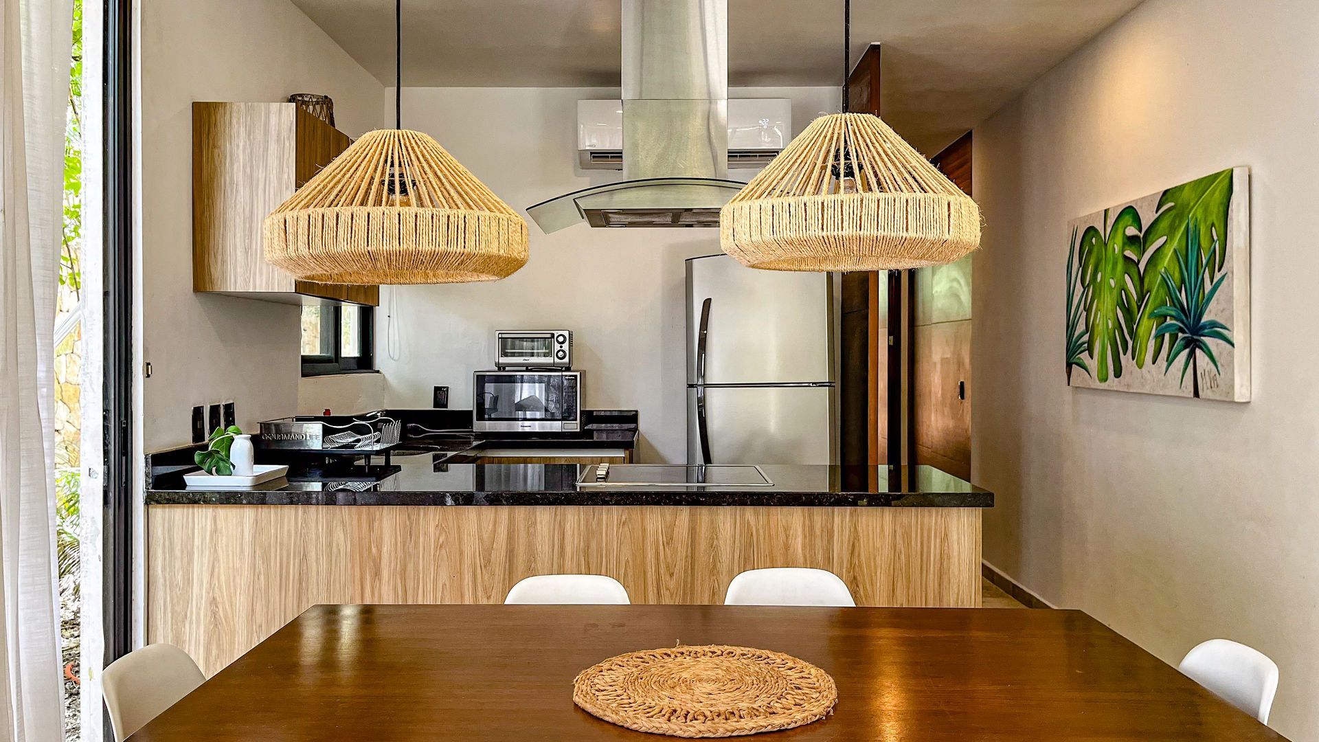 Cocina con comedor; gabinetes de madera, encimera negra, luces colgantes tejidas, obras de arte y una mesa con sillas.