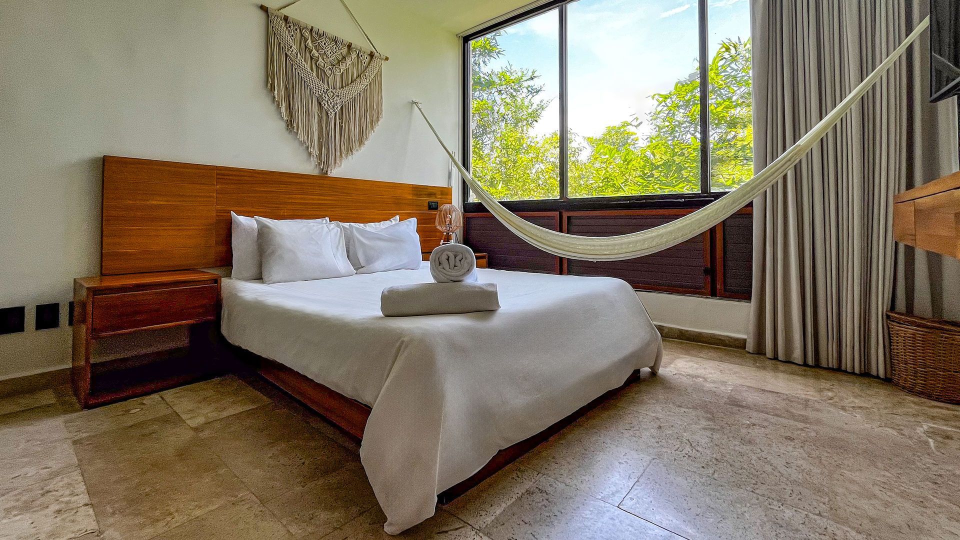 Dormitorio con cama, mesita de noche, hamaca y un gran ventanal con vista a los árboles. Predominan los colores neutros.