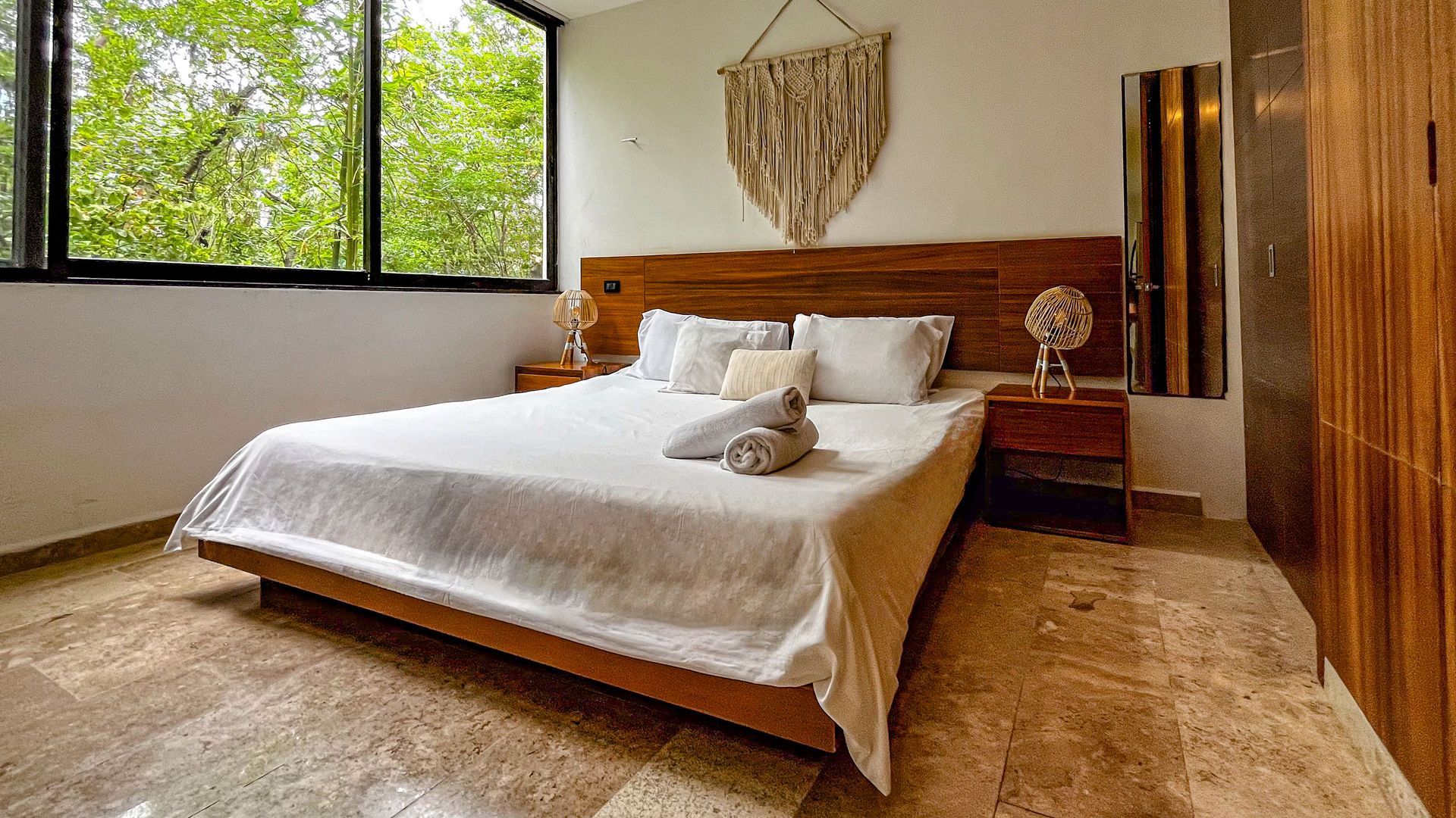 Dormitorio con cama grande, cabecero de madera y ventana con vistas a los árboles.