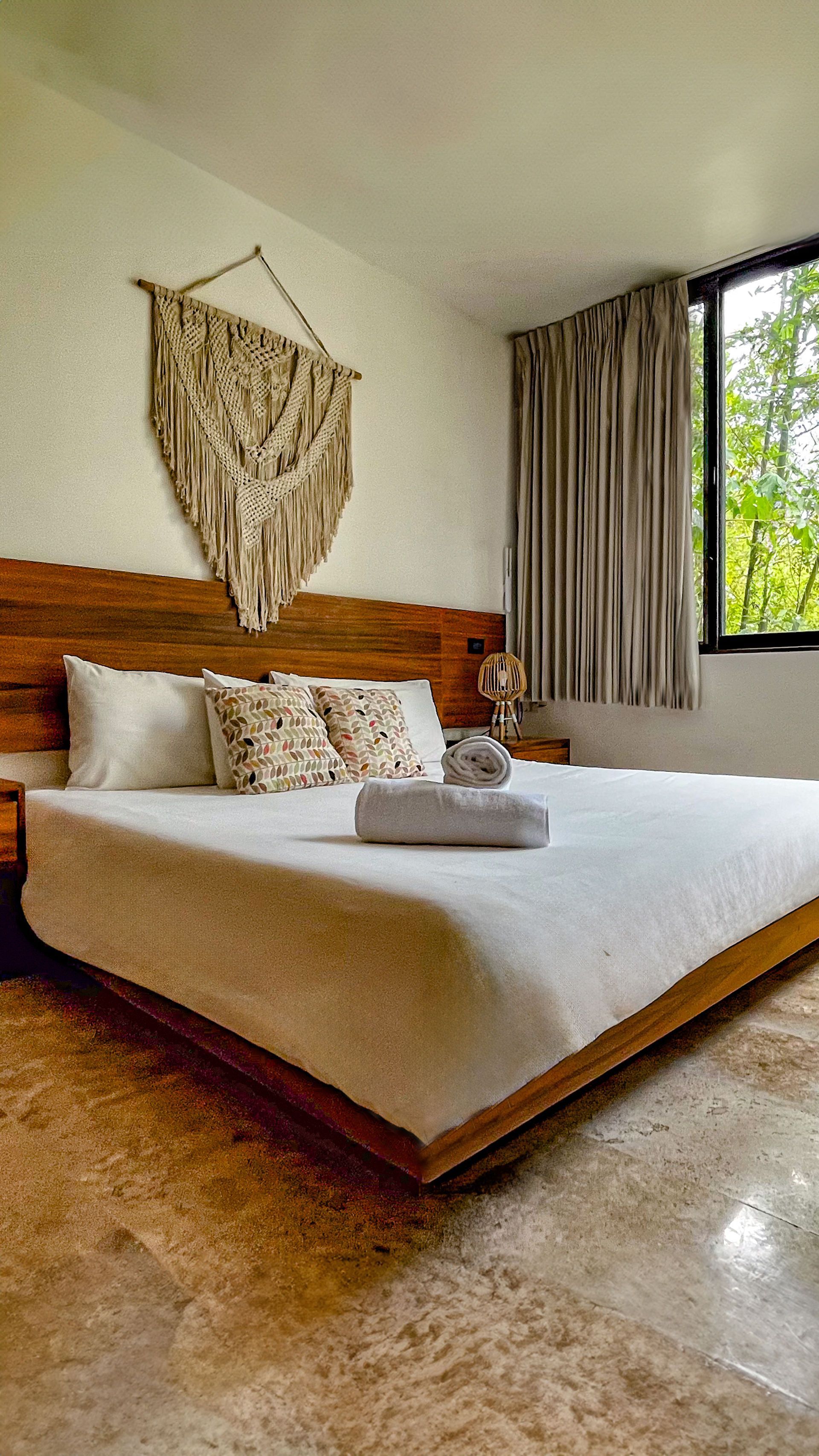 Dormitorio con estructura de cama de madera, ropa de cama blanca, tapiz de macramé y ventana con cortinas.