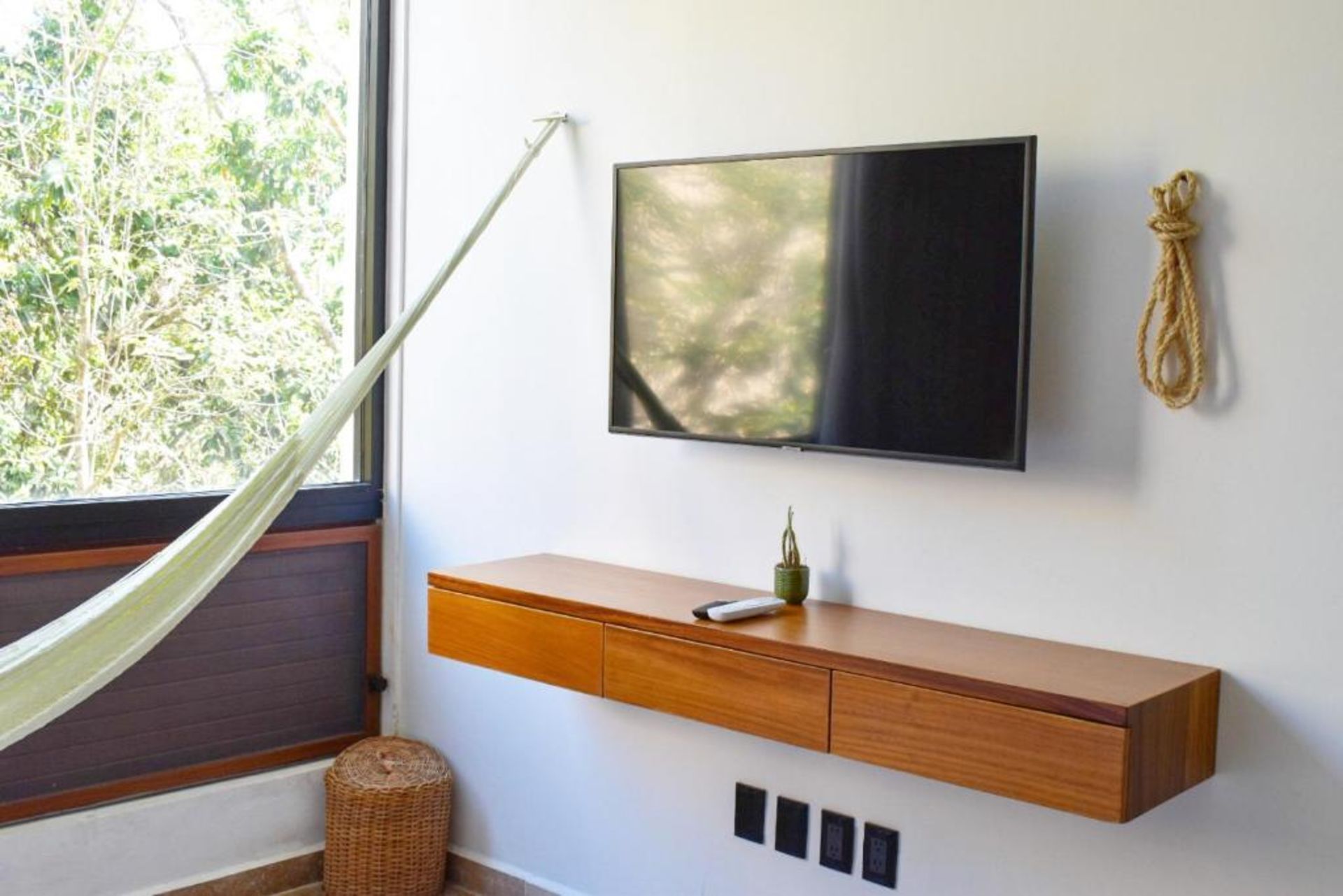 Un televisor montado en la pared sobre una consola flotante de madera con una hamaca y una ventana.