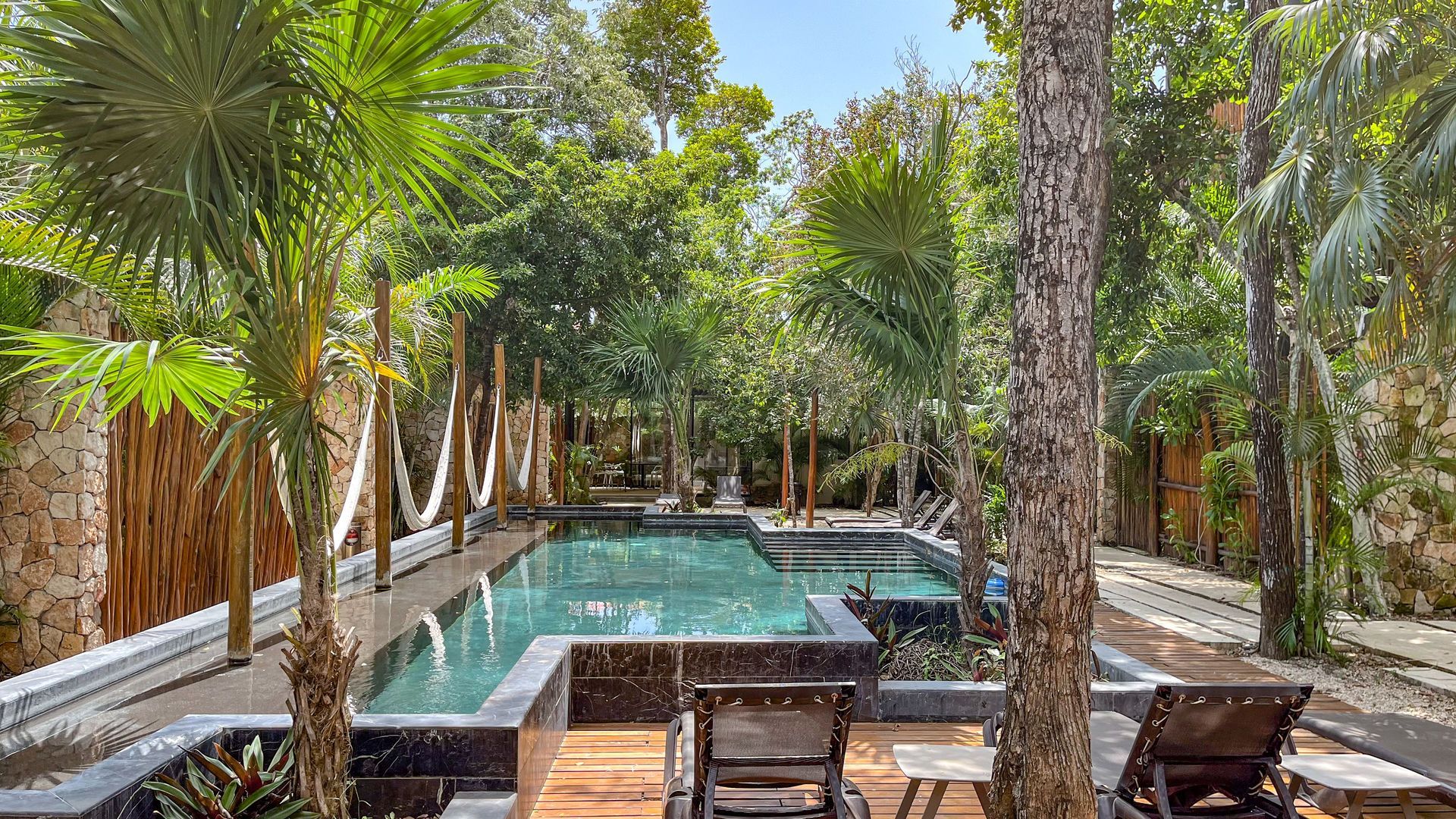 Piscina rodeada de exuberante vegetación y hamacas; dos sillas frente a la piscina.