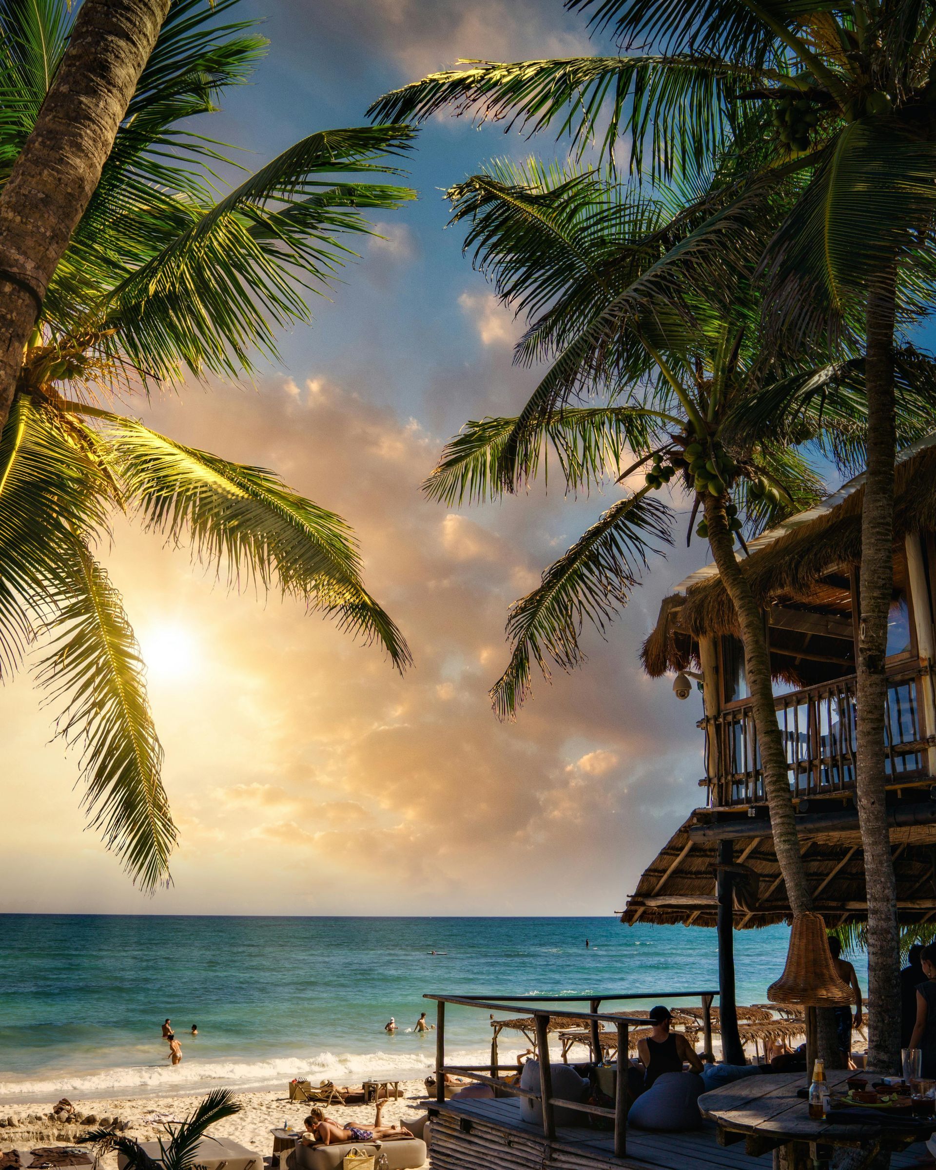 Escena de playa con palmeras, océano, cabaña de madera y cielo al atardecer.