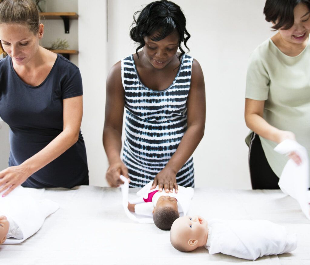 Three people folding white towels beside baby dolls on a table in a bright room