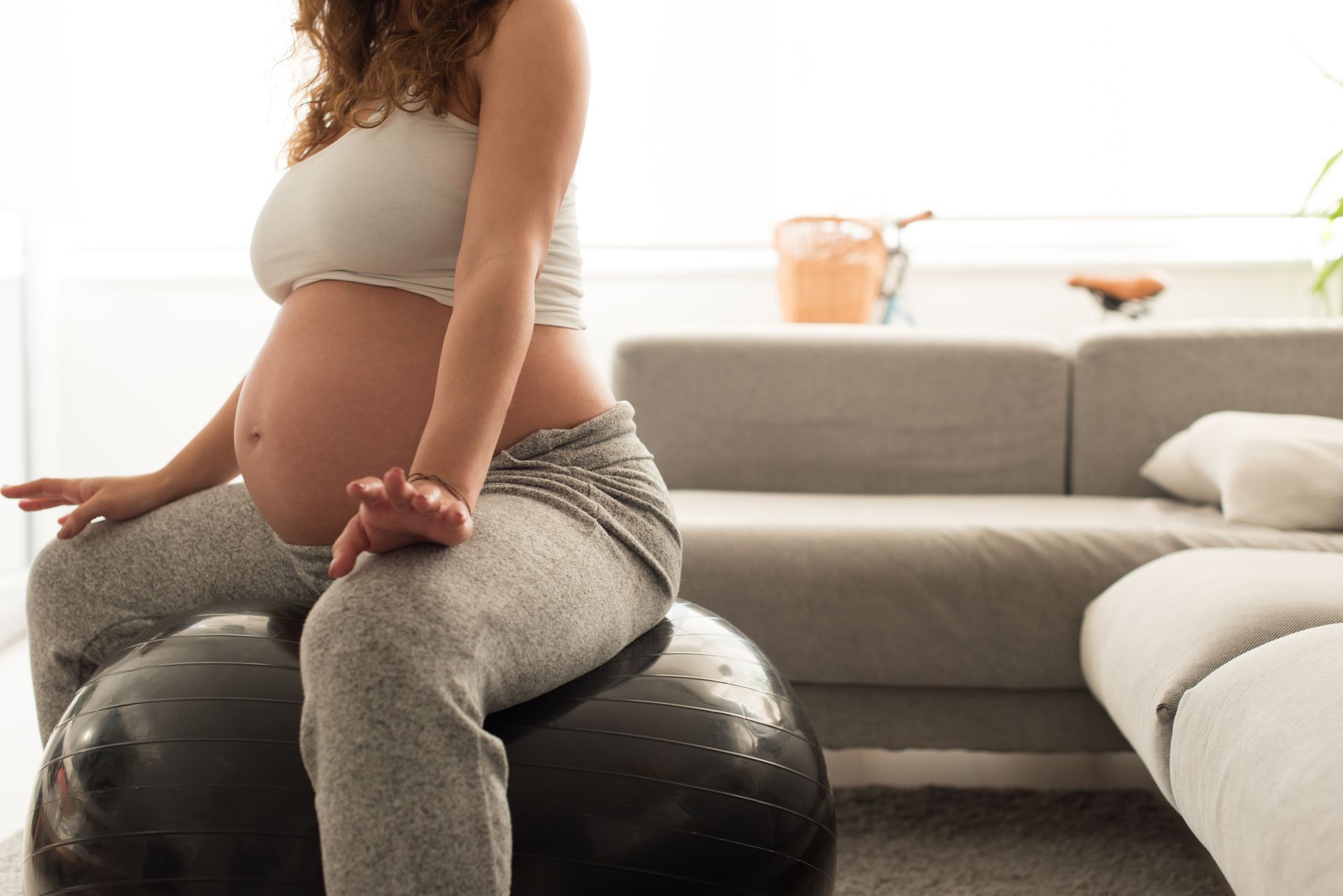 Pregnant person sitting cross-legged on a black exercise ball in a bright living room