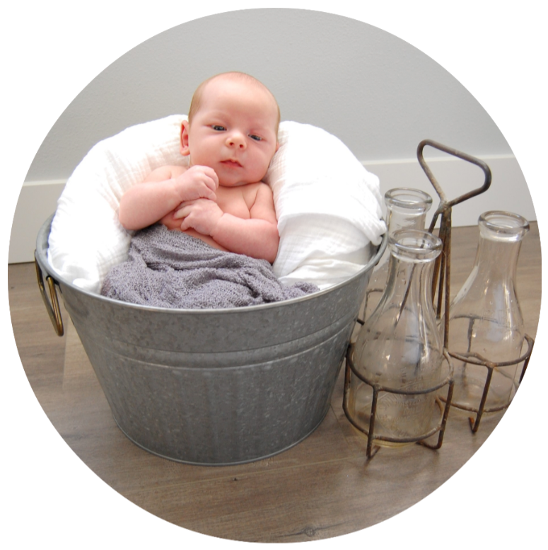 Baby resting in a metal tub with white pillows, beside two glass bottles on a wooden floor.