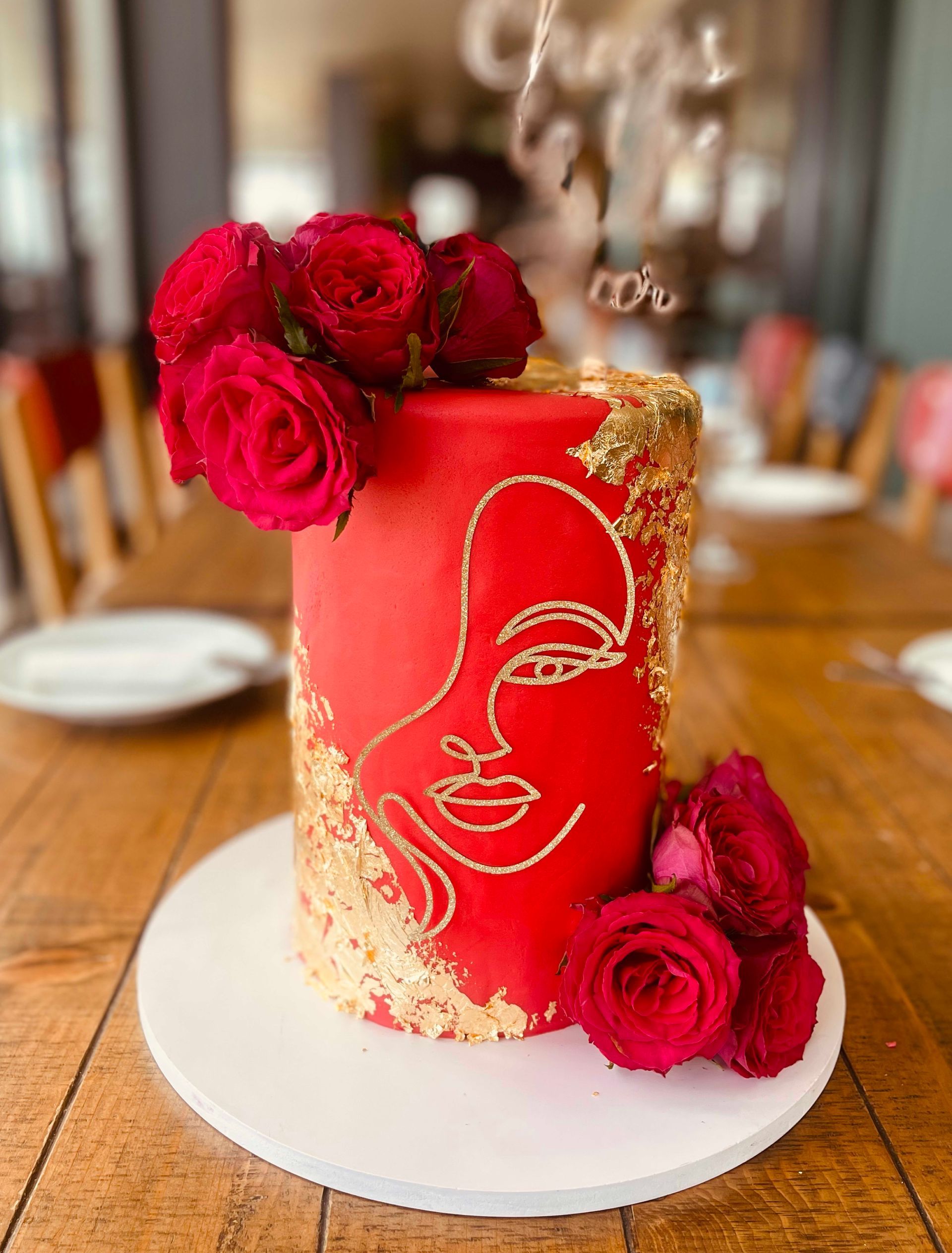 Red cake with gold face design, roses, and gold leaf on white plate. Wooden table setting.