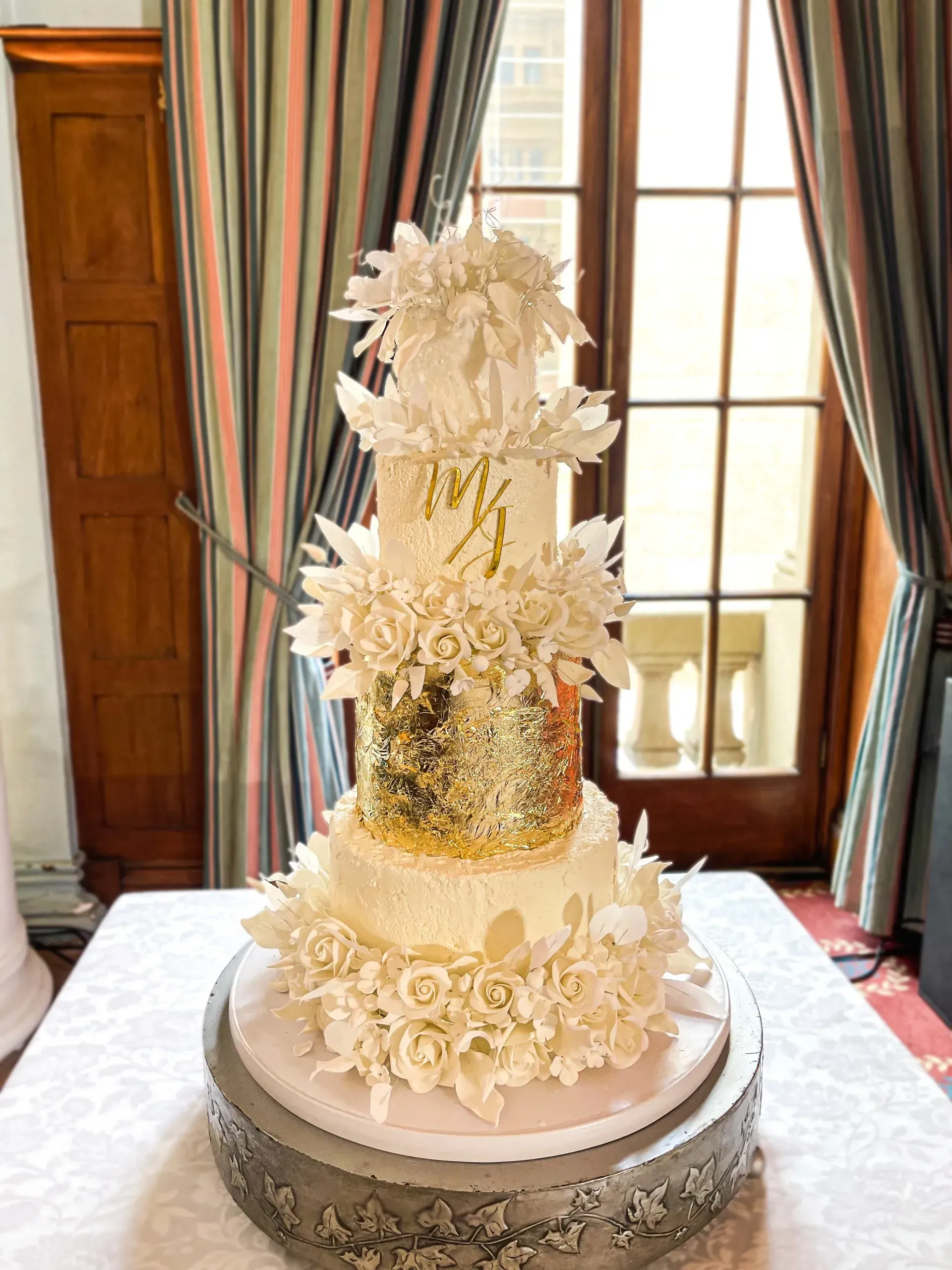 Elegant white wedding cake with gold accents and floral decorations on a pedestal table, in a room with a window.