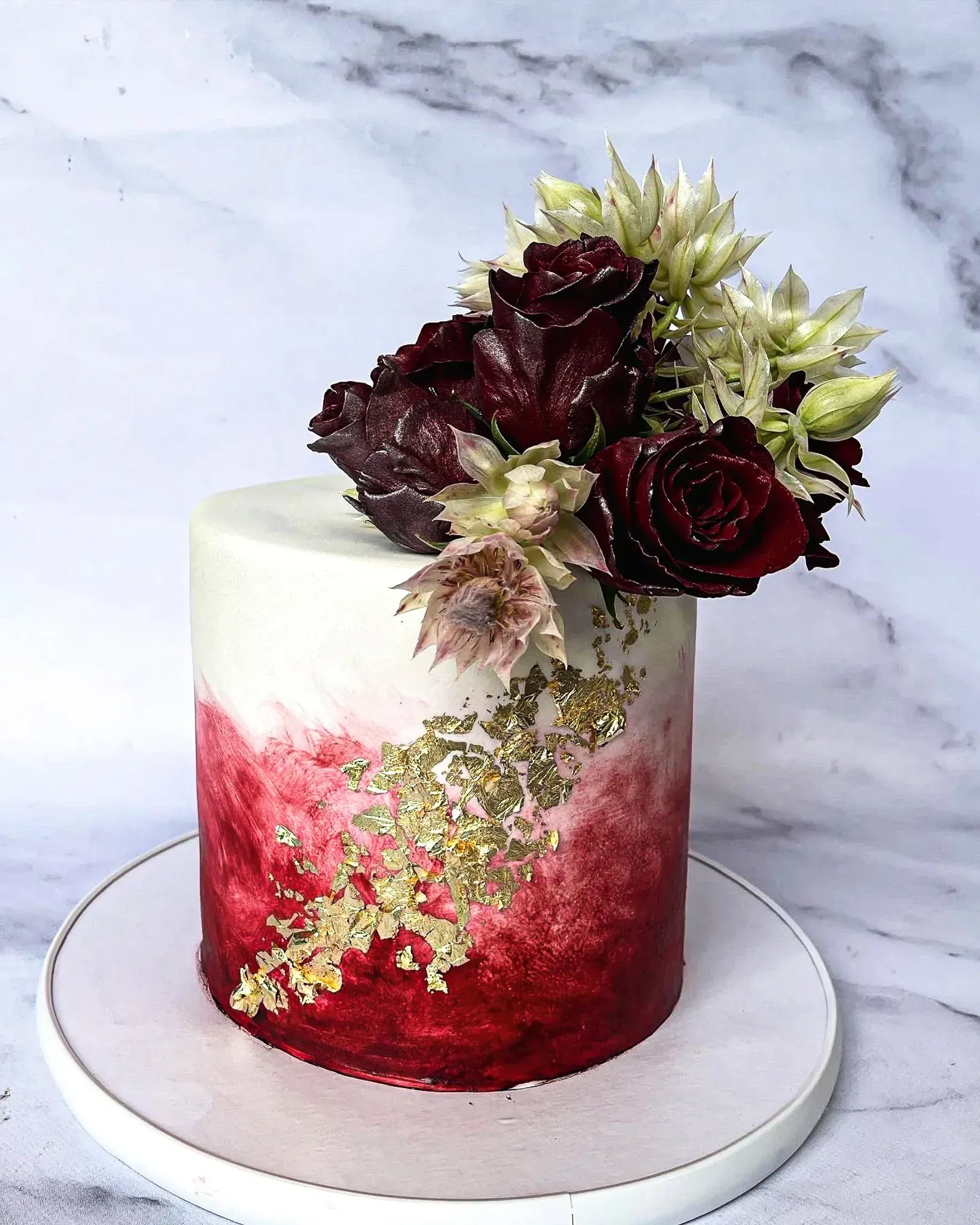 Cake with red ombre frosting, gold leaf accents, and floral arrangement on a white plate.