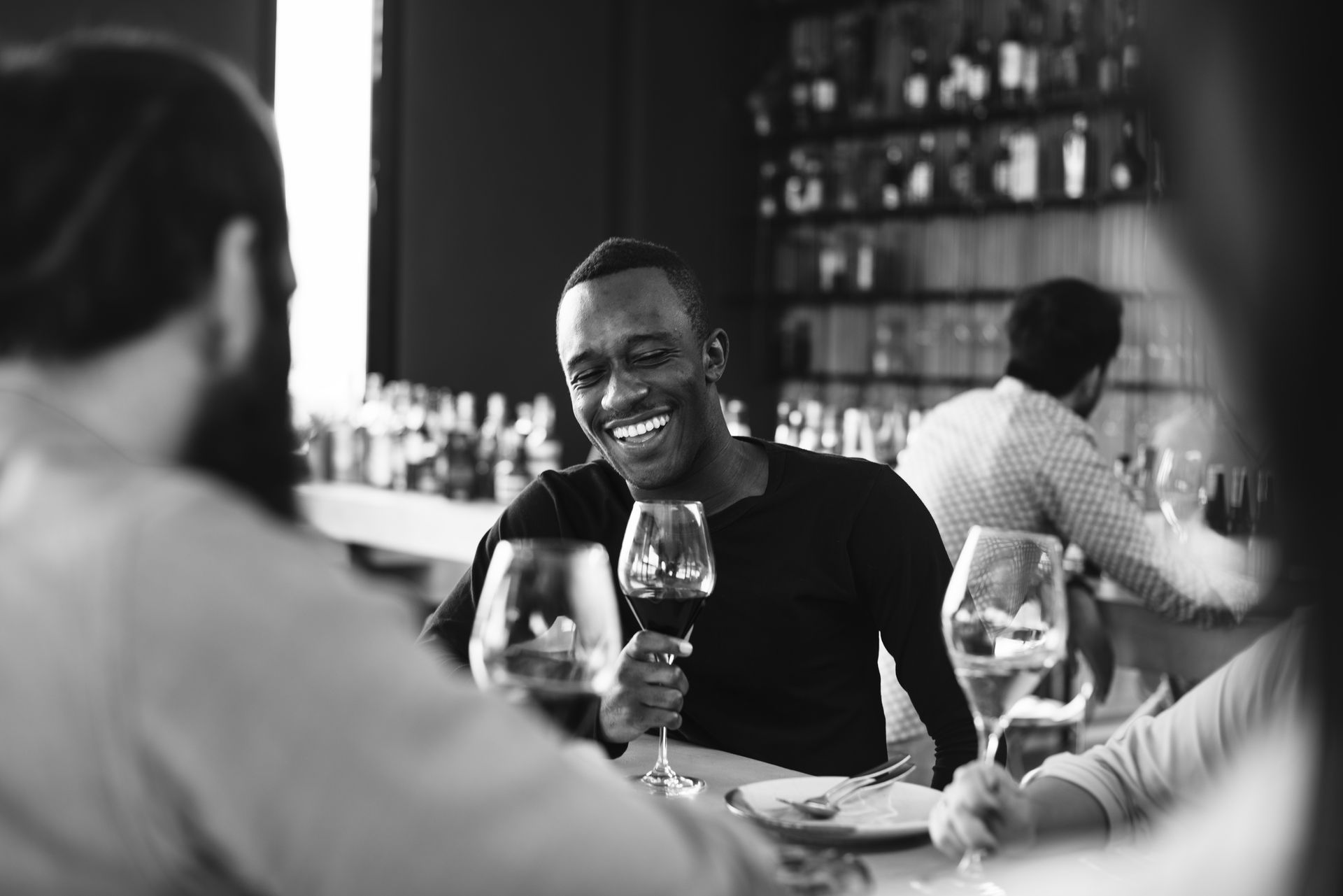 Black and white photo of a smiling person holding a wine glass at a restaurant table with others.