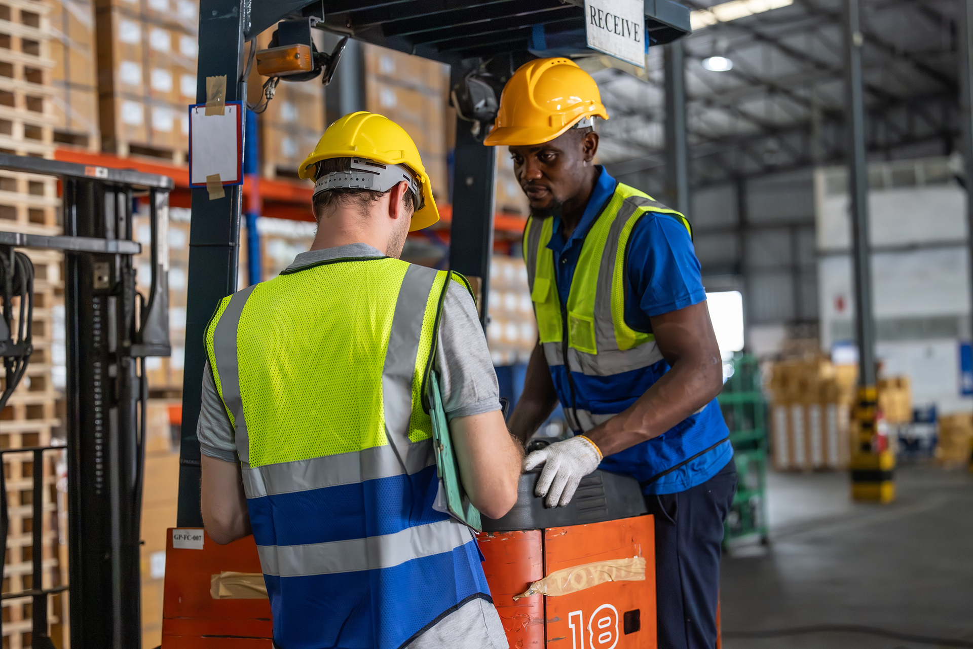 Two workers in hard hats and safety vests at a warehouse near a forklift.