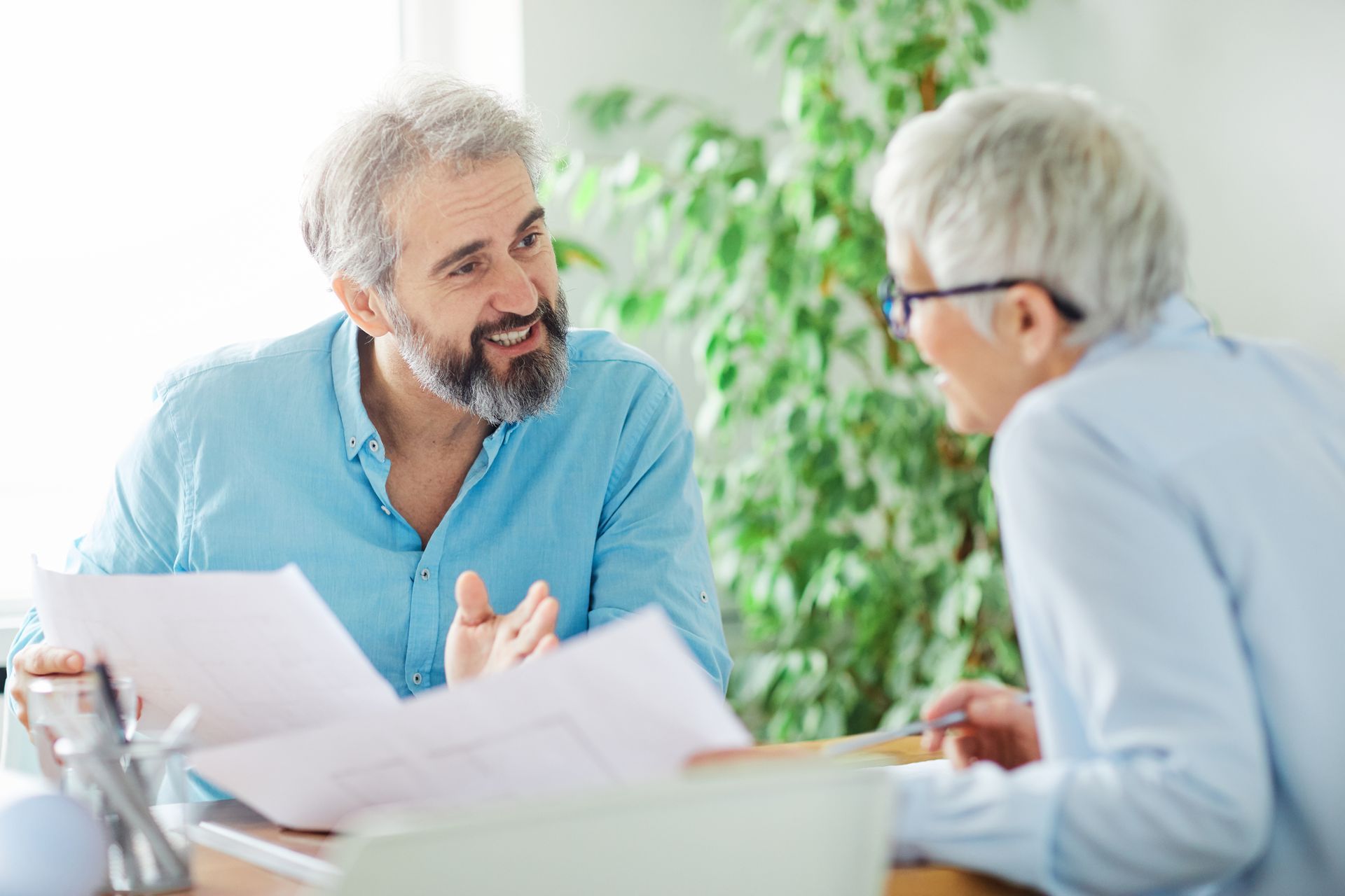 Man with beard gestures, discussing papers with a woman wearing glasses, near a plant.