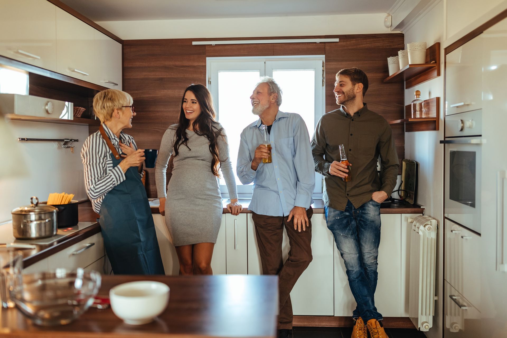 Four people in a kitchen talking: woman in apron, pregnant woman, older man, younger man.