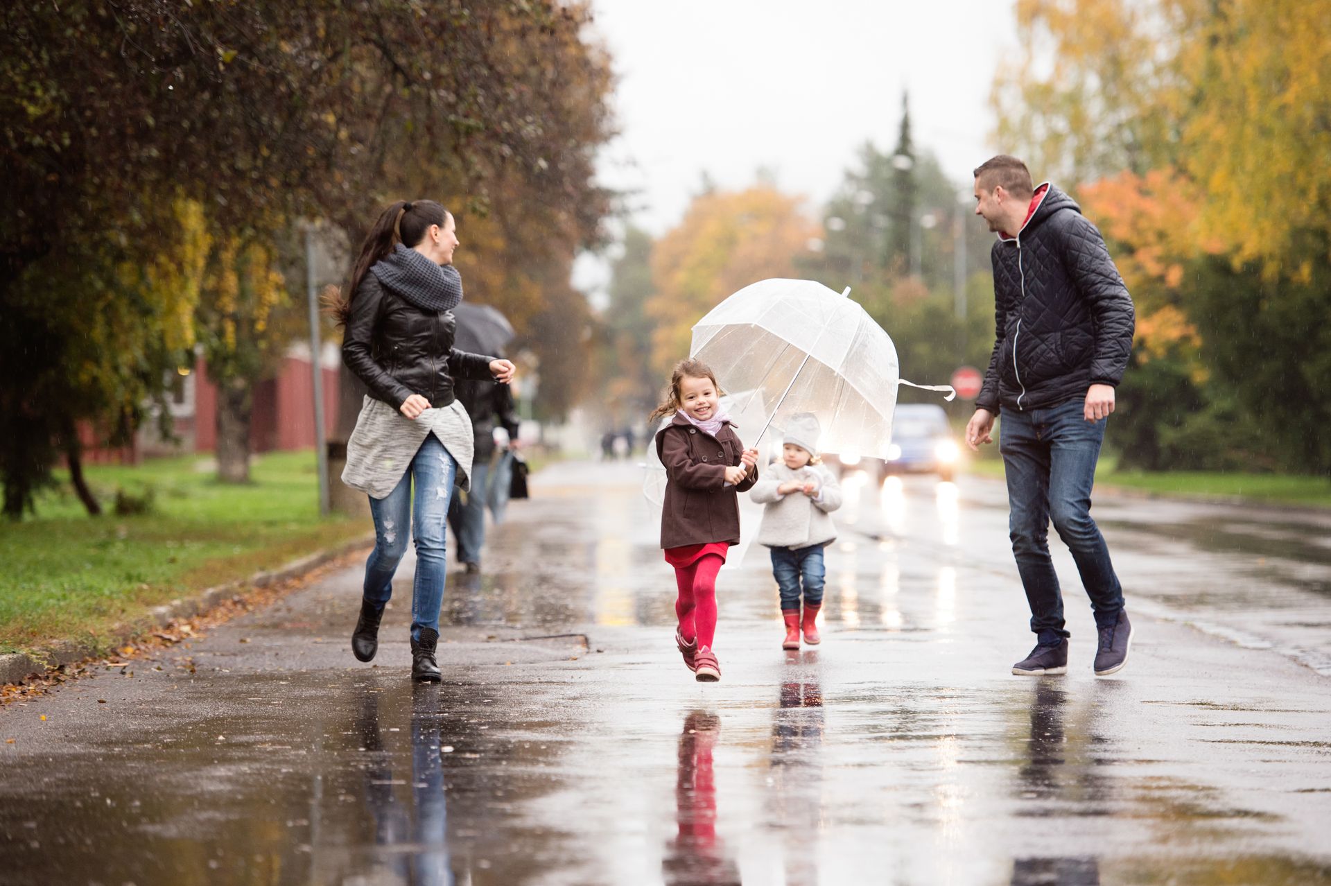 Family of four running and laughing in the rain on a street. One holds an umbrella. Fall foliage in background.