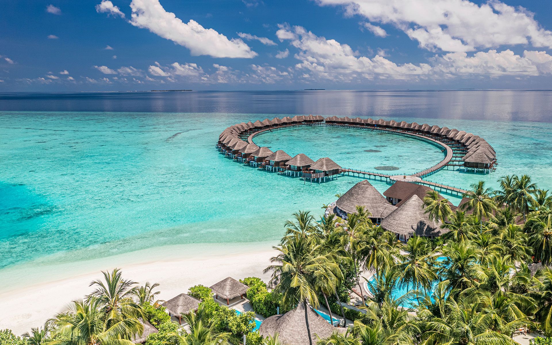 Overwater bungalows in a lagoon; turquoise water, white sand beach, palm trees, blue sky.