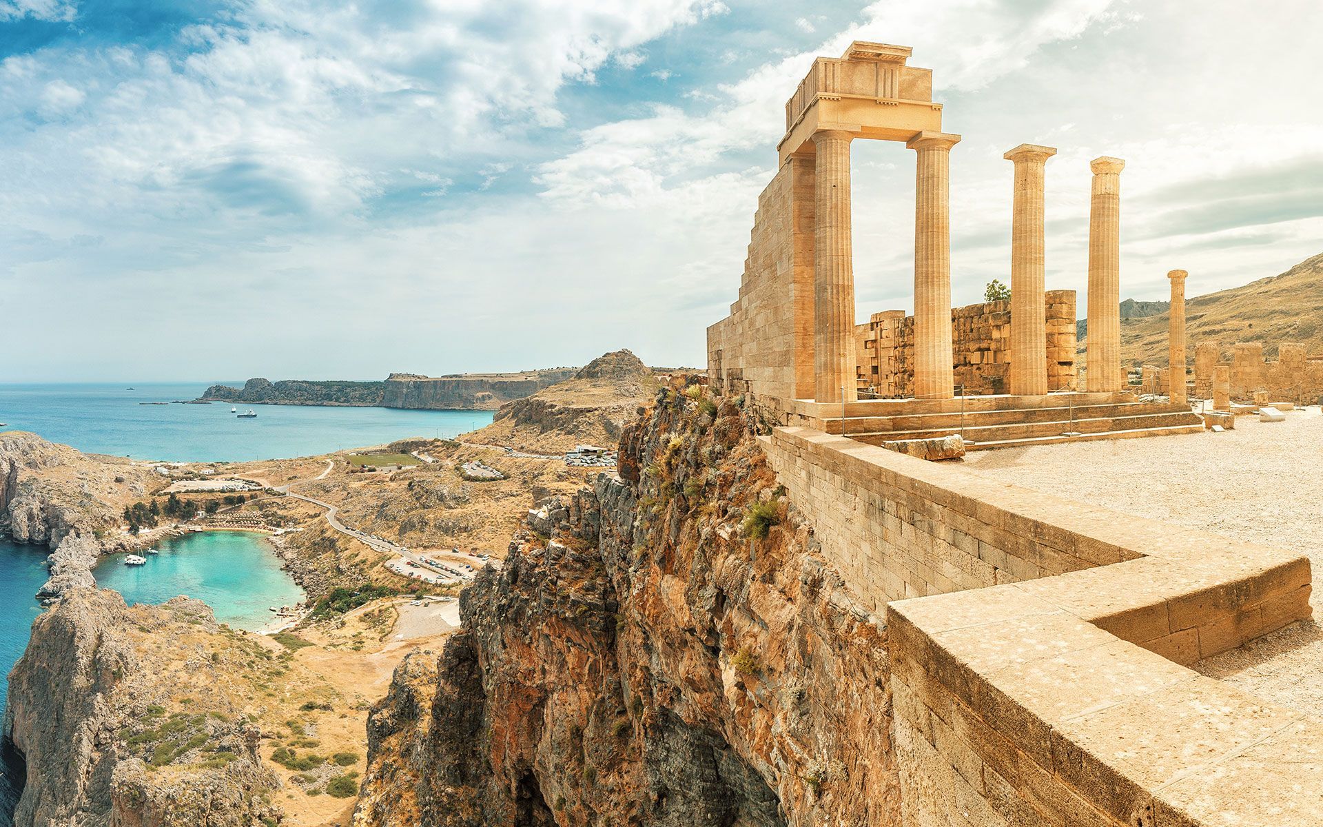 Ruins atop cliff overlooking a Greek coastal town with blue water and sky.