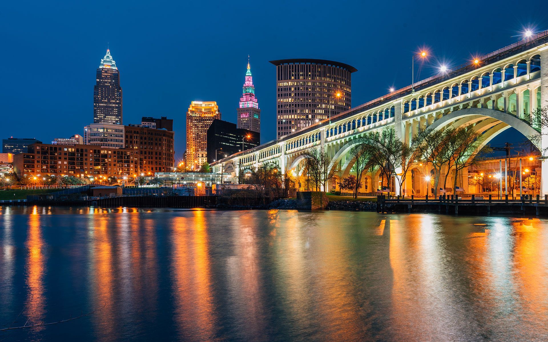 Nighttime cityscape of Cleveland with bridge, buildings, and water reflecting lights.