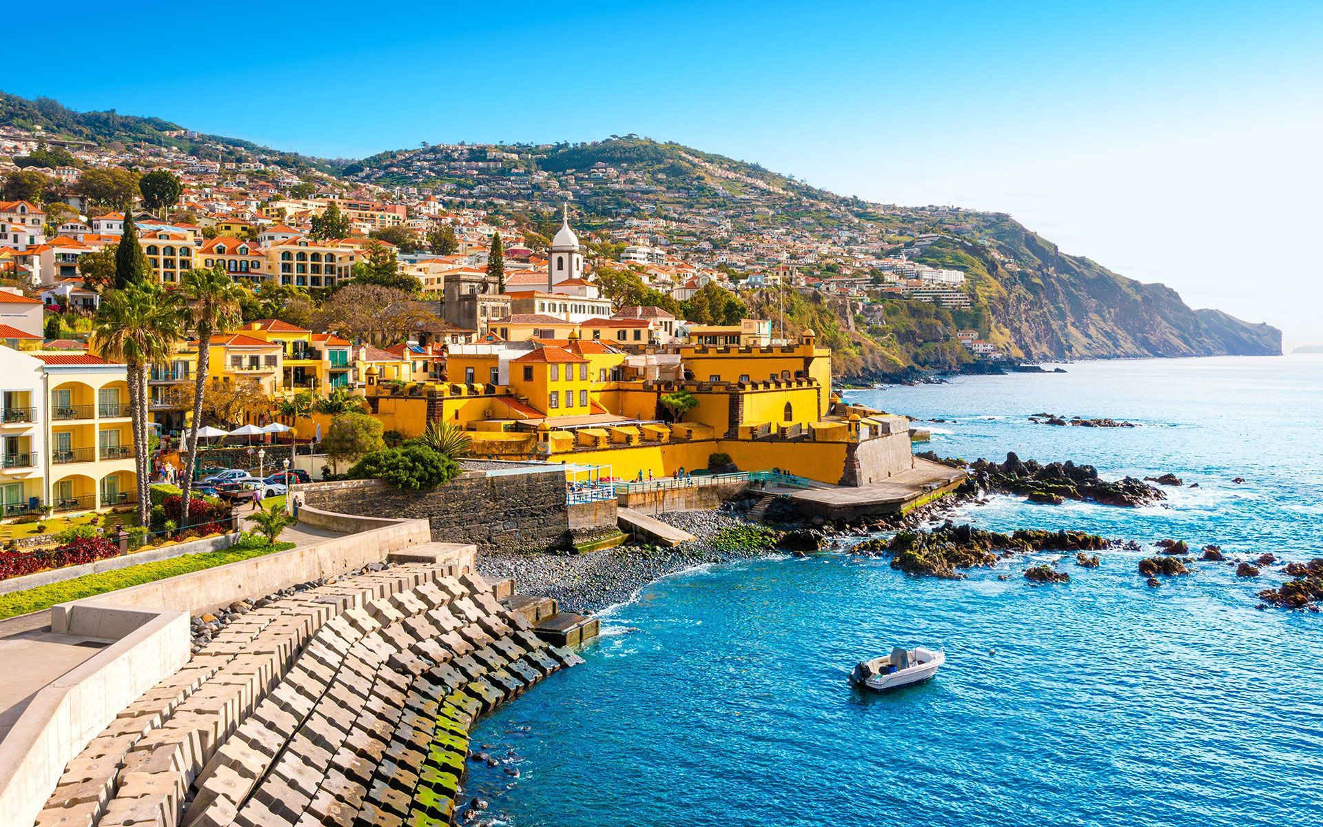 Coastal city in Madeira, Portugal, with yellow buildings, blue sea, and green hillside under a bright blue sky.