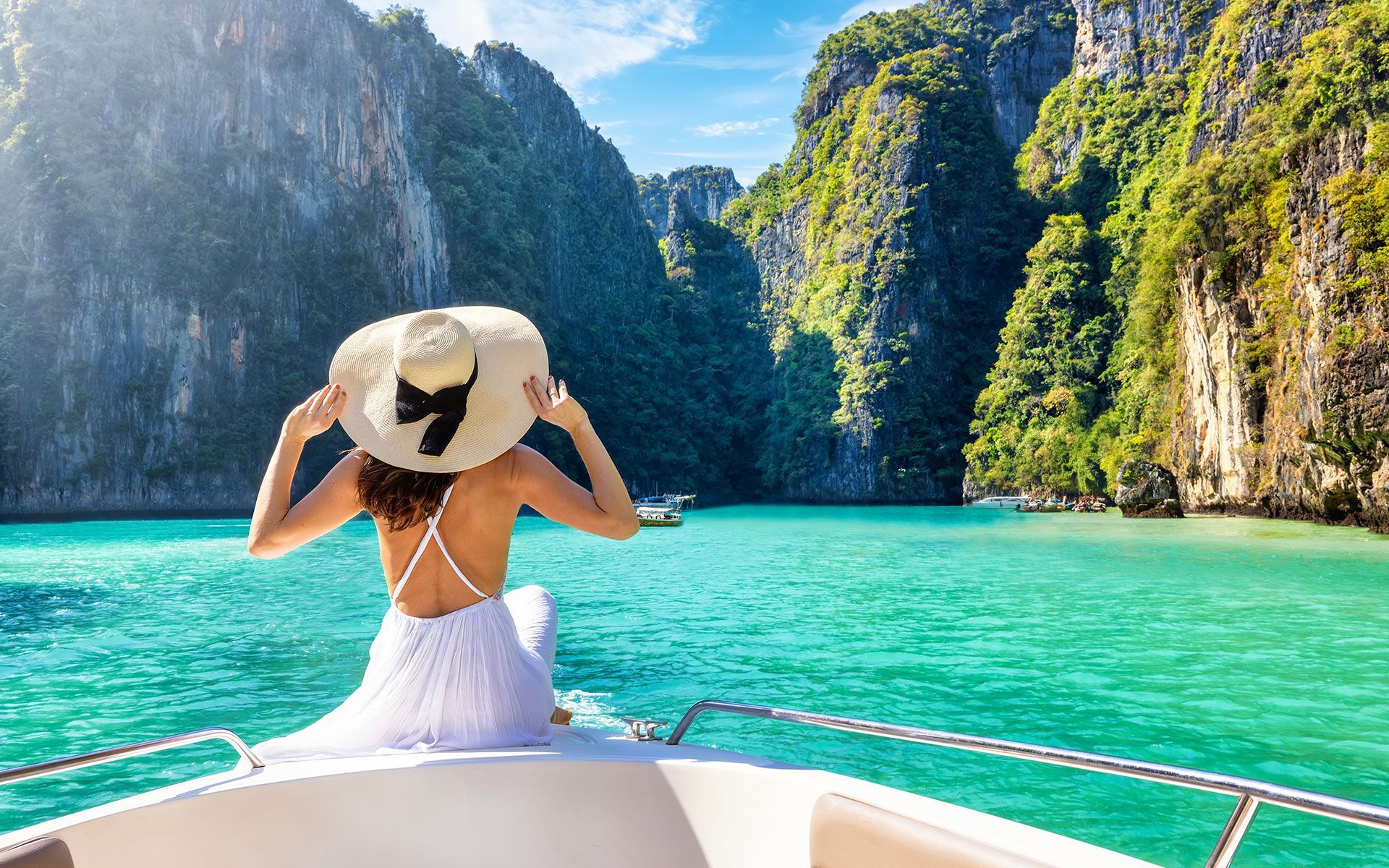 Woman in white dress and hat on boat, admiring turquoise water and lush cliffs.