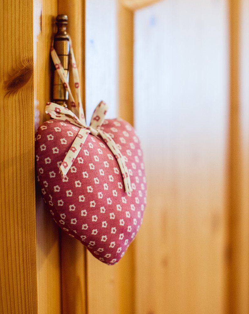 Ein rosa gemustertes, herzförmiges Säckchen hängt mit einer Schleife an einem Türgriff aus Holz.