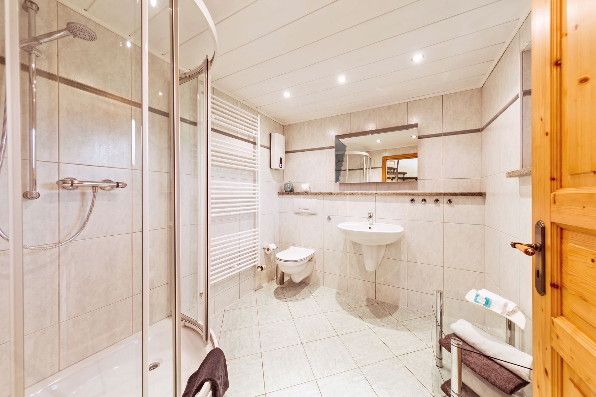 Helles, gefliestes Badezimmer mit Dusche, Waschbecken, Toilette, Spiegel und Holztürdetails