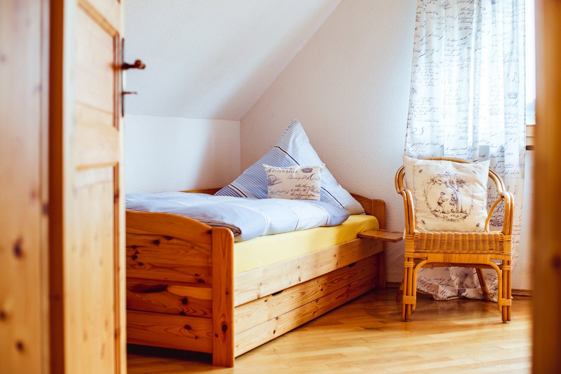 Gemütliches Dachgeschossschlafzimmer mit hölzernem Tagesbett, Korbsessel und Sonnenlicht durch ein mit Spitzengardinen behängtes Fenster