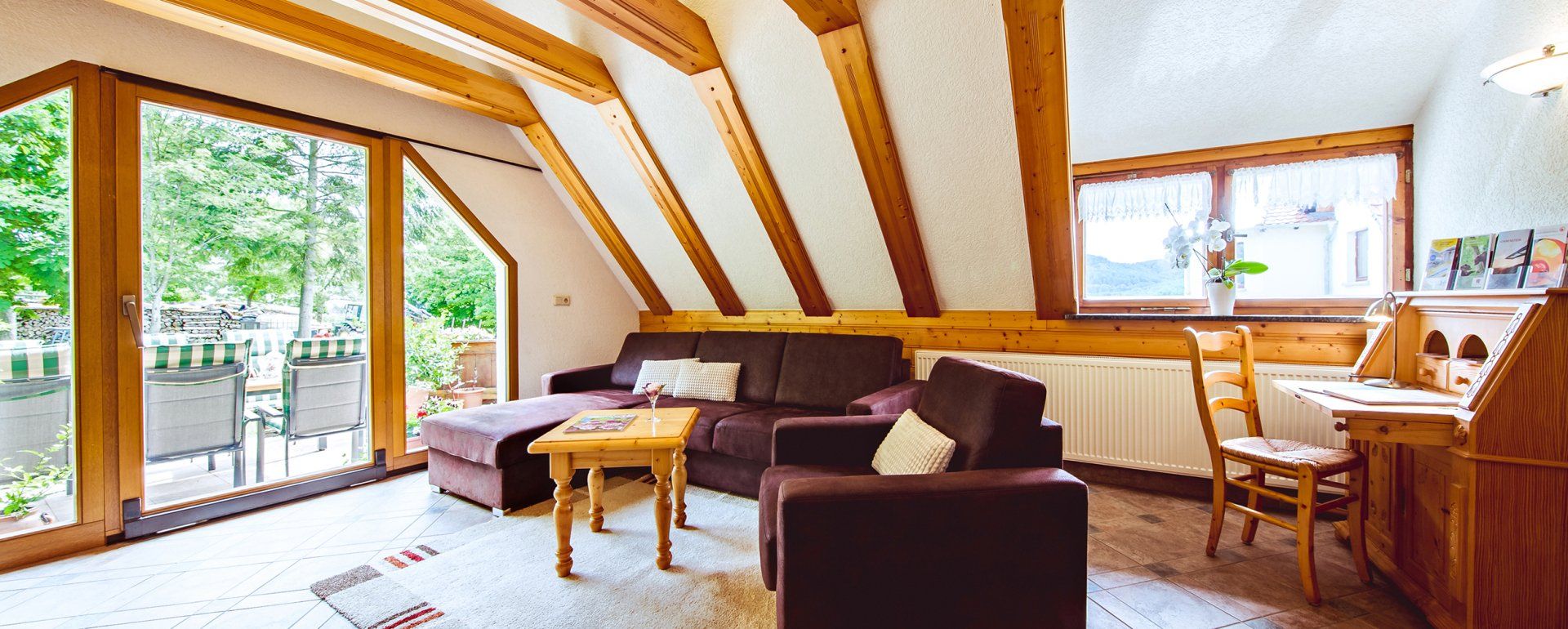 Helles Wohnzimmer mit sichtbaren Holzbalken, braunem Sofa, Sessel, Couchtisch und Balkonblick