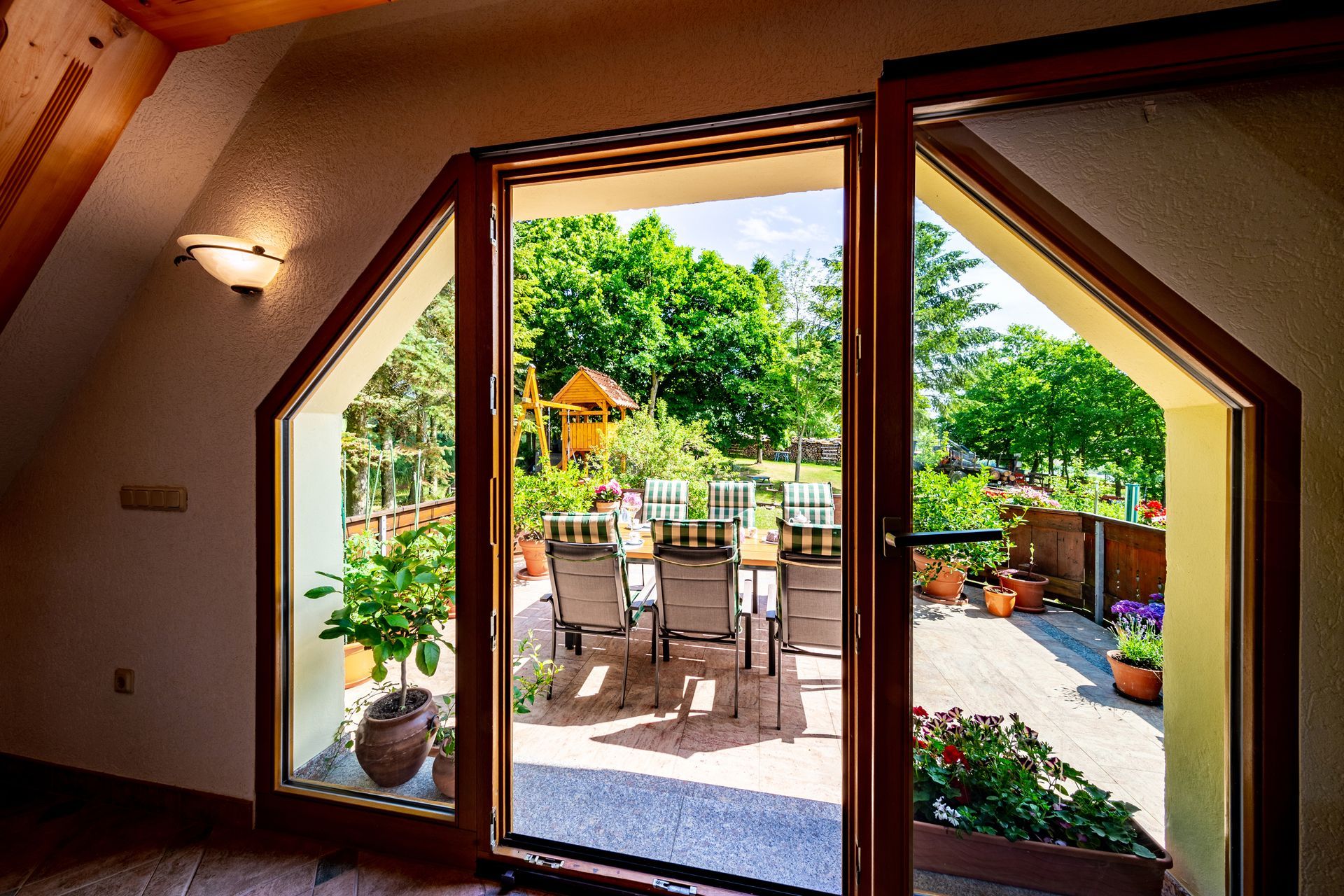 Blick durch Glastüren auf eine sonnige Terrasse mit Tisch, Stühlen und üppigem Grün.