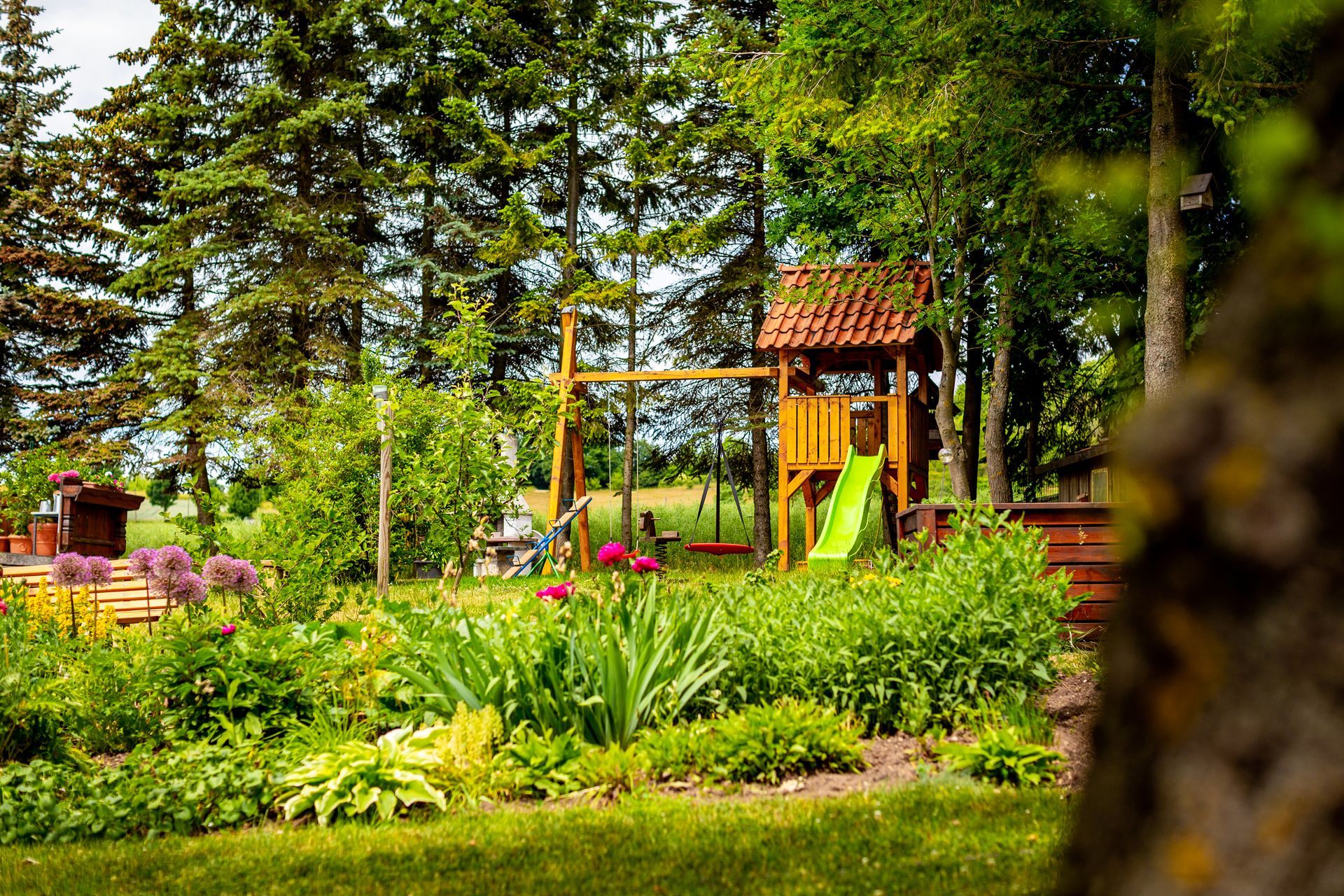 Ein üppiger Garten mit bunten Blumen, Bäumen und einem kleinen Holzspielhaus mit gelber Rutsche.