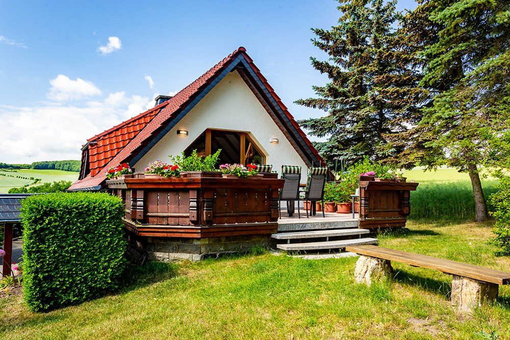Kleines Häuschen mit rotem Dach, Holzterrasse und Treppe, gelegen auf einem sonnigen, grasbewachsenen Feld.