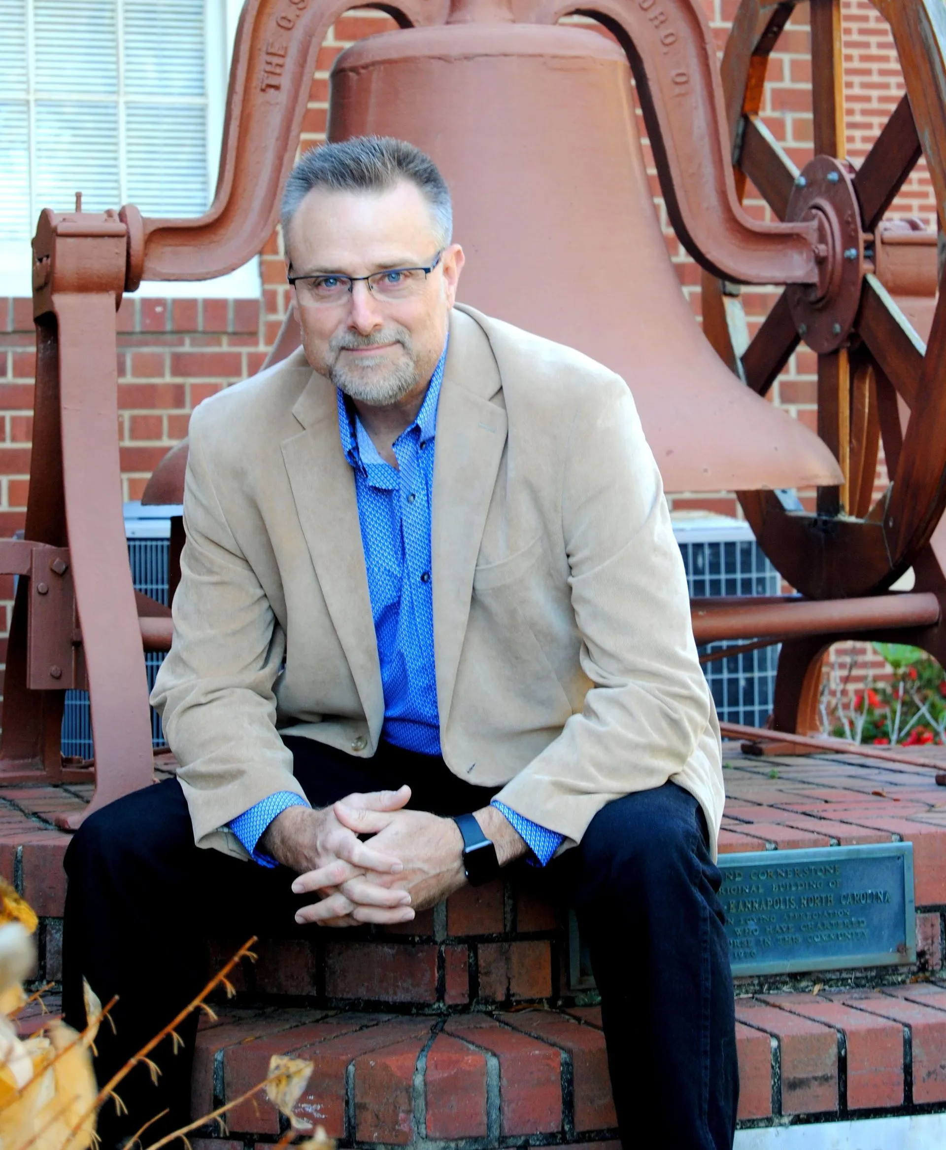 A man in a tan jacket is sitting in front of a large bell