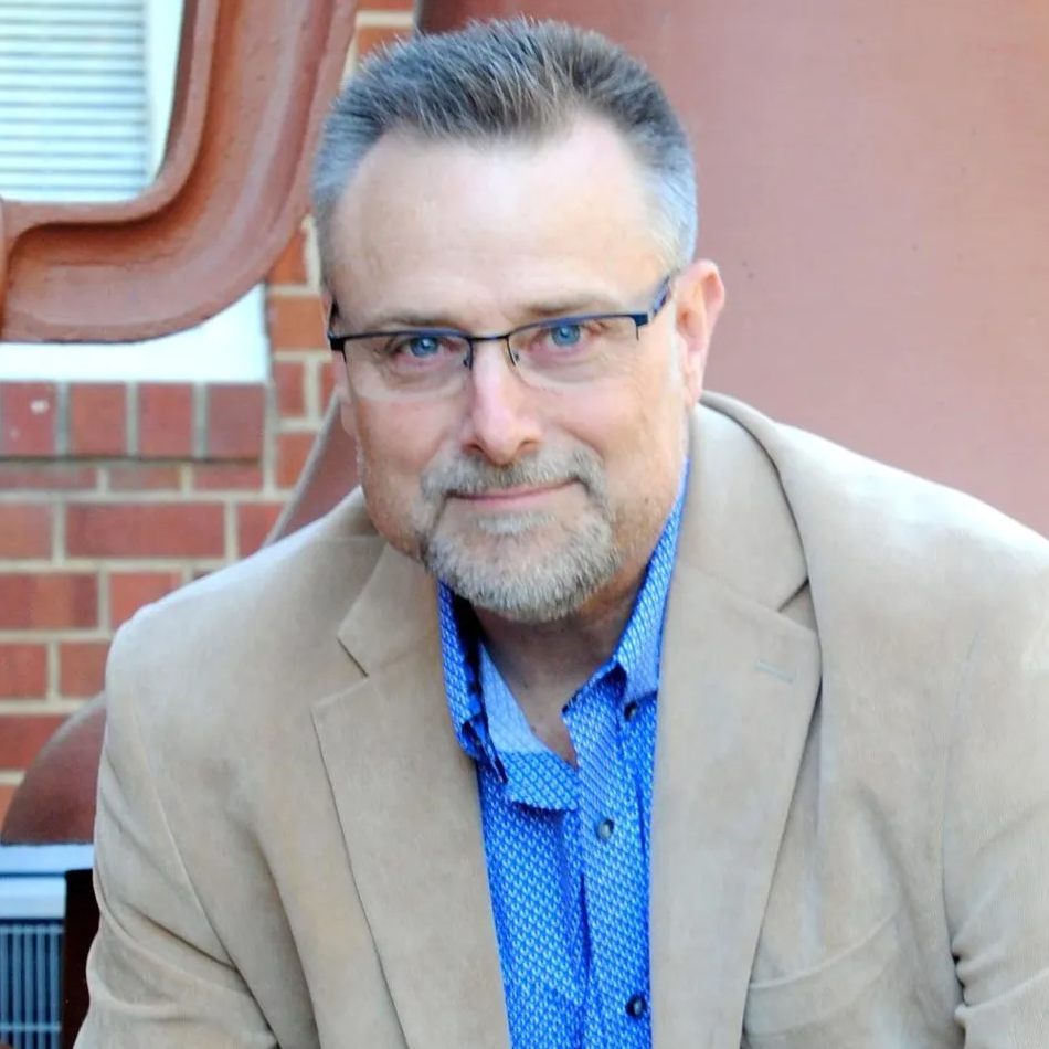 A man wearing glasses and a tan jacket is sitting in front of a brick wall