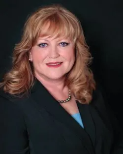 A woman in a black jacket and blue shirt is smiling for the camera.