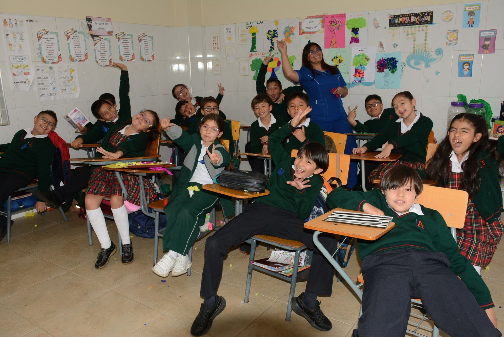 Aula llena de alumnos sonriendo y levantando la mano, con coloridos carteles en la pared.