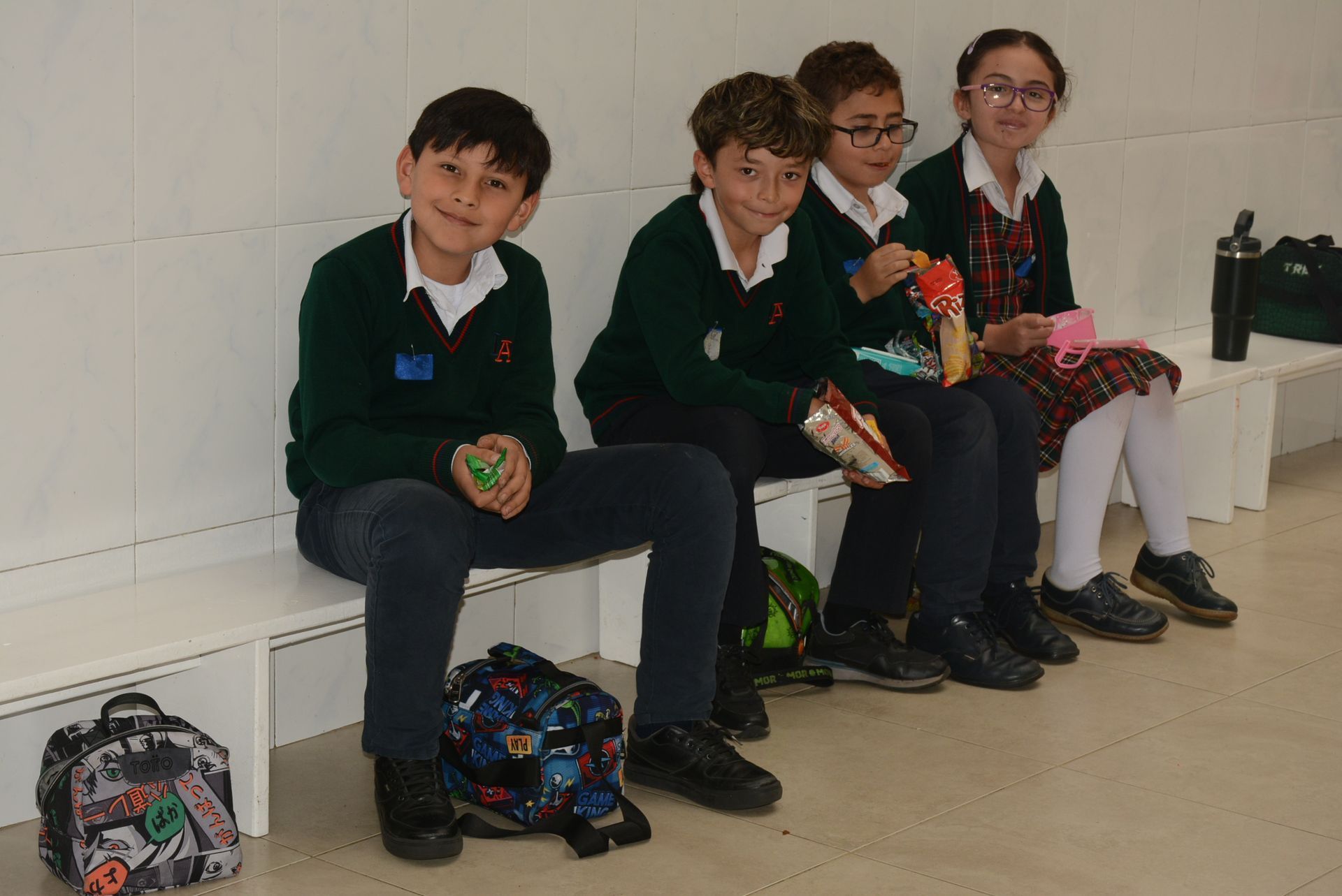Cuatro estudiantes con uniformes verdes están sentados en un banco en un pasillo, con sus mochilas en el suelo frente a ellos.