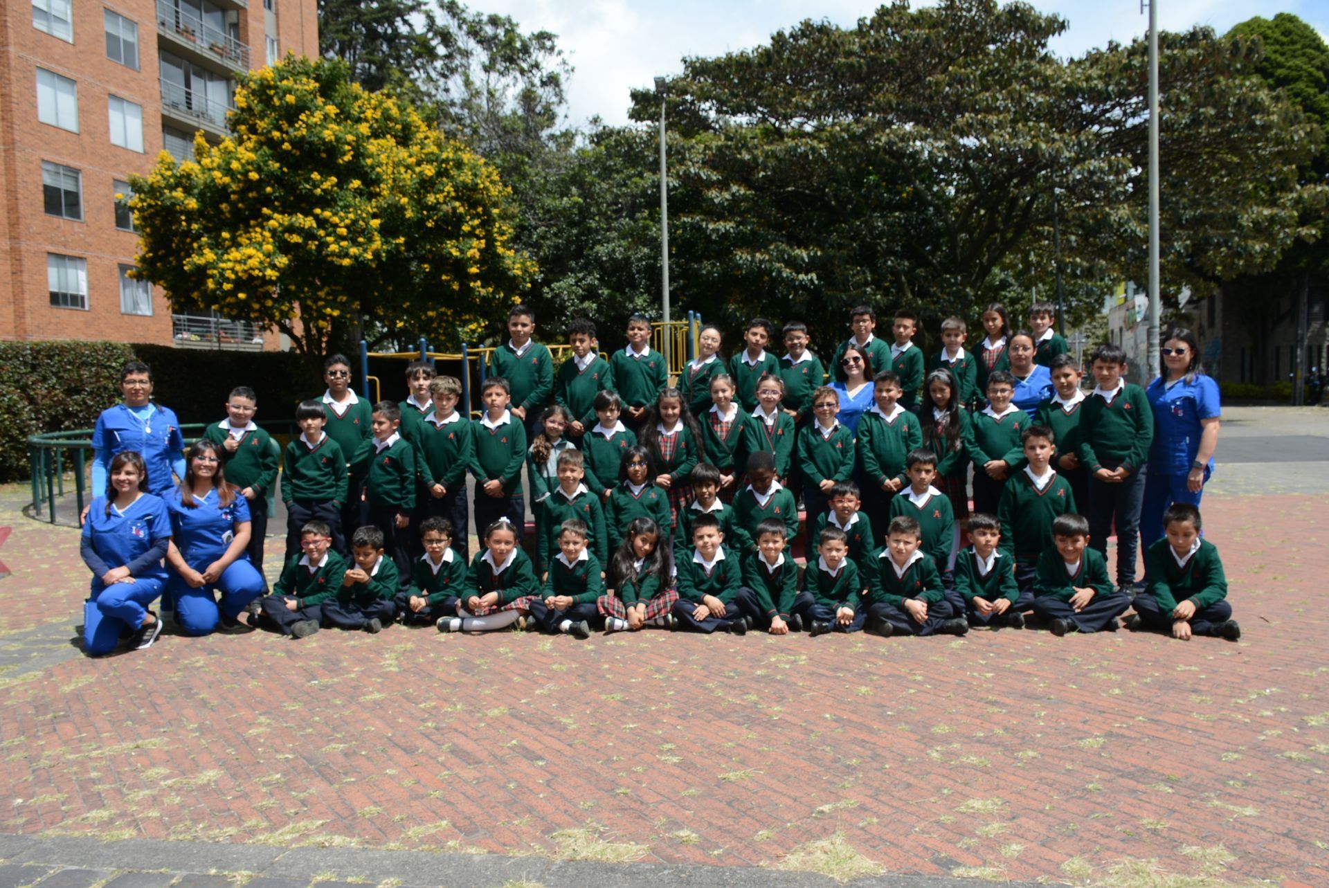 Un grupo numeroso de personas con uniformes deportivos verdes y azules posa al aire libre frente a un edificio y árboles.