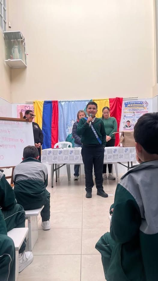 Dos personas hablando en un pequeño evento en un espacio cerrado, con coloridas banderas y público sentado al fondo.