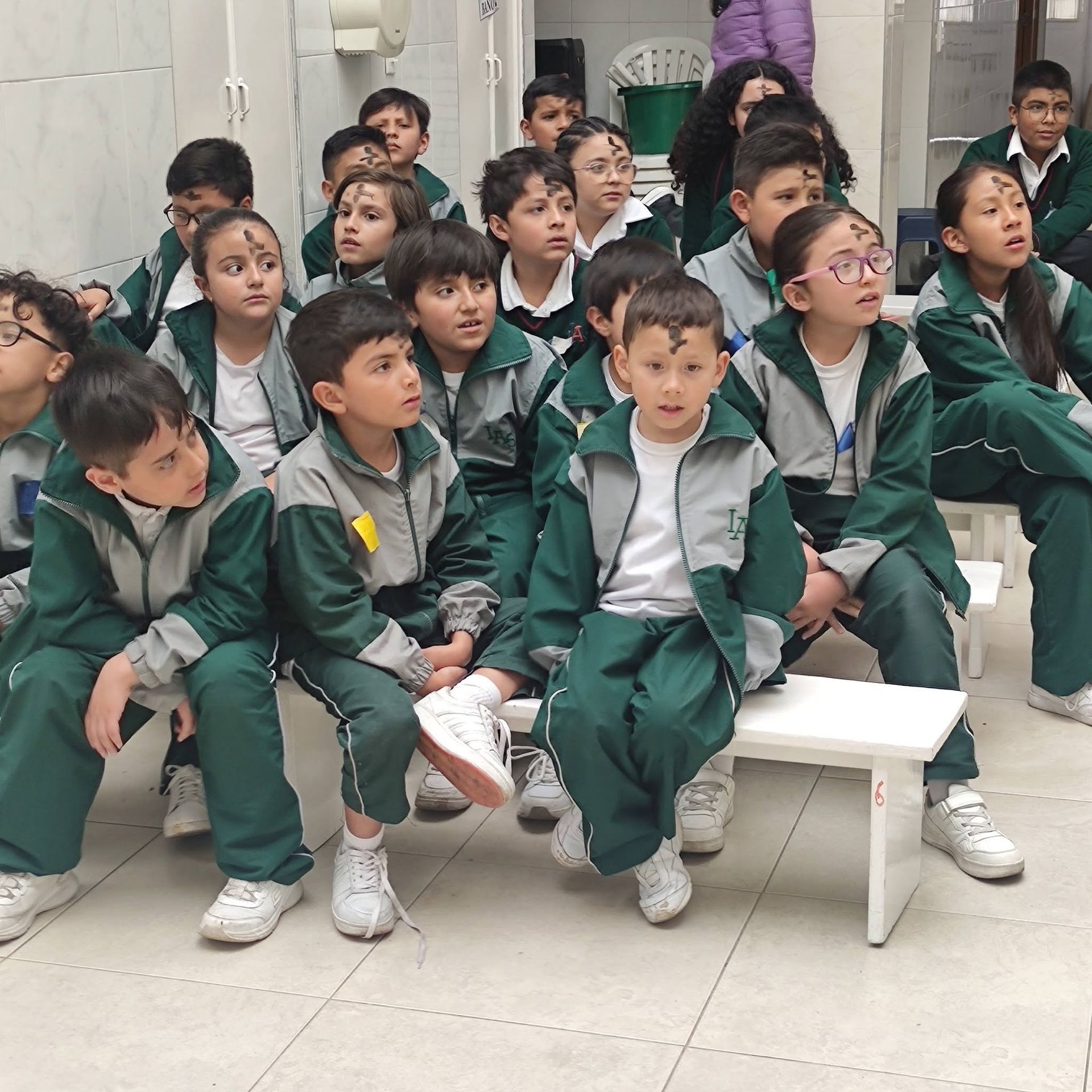 Estudiantes con uniformes verdes sentados en el interior, mirando atentamente hacia arriba en un aula o área de asamblea.