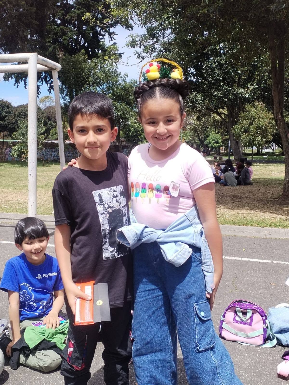 Dos niños de pie en un parque, uno con una camiseta negra y el otro con una camiseta rosa y una chaqueta vaquera, con mochilas cerca.