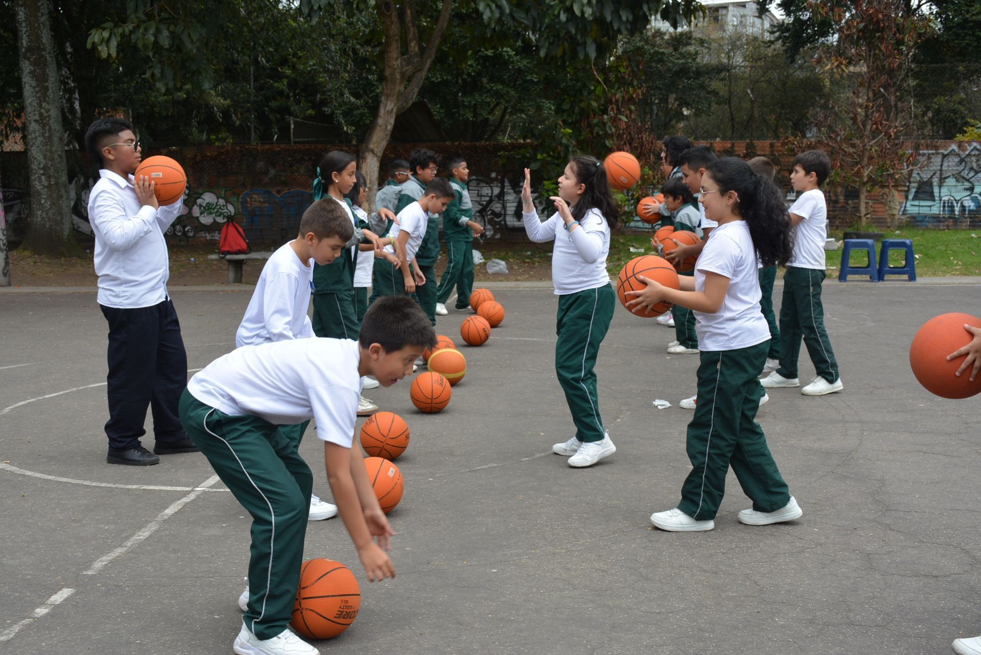 Estudiantes practicando el regate de baloncesto en una cancha al aire libre con pelotas naranjas.
