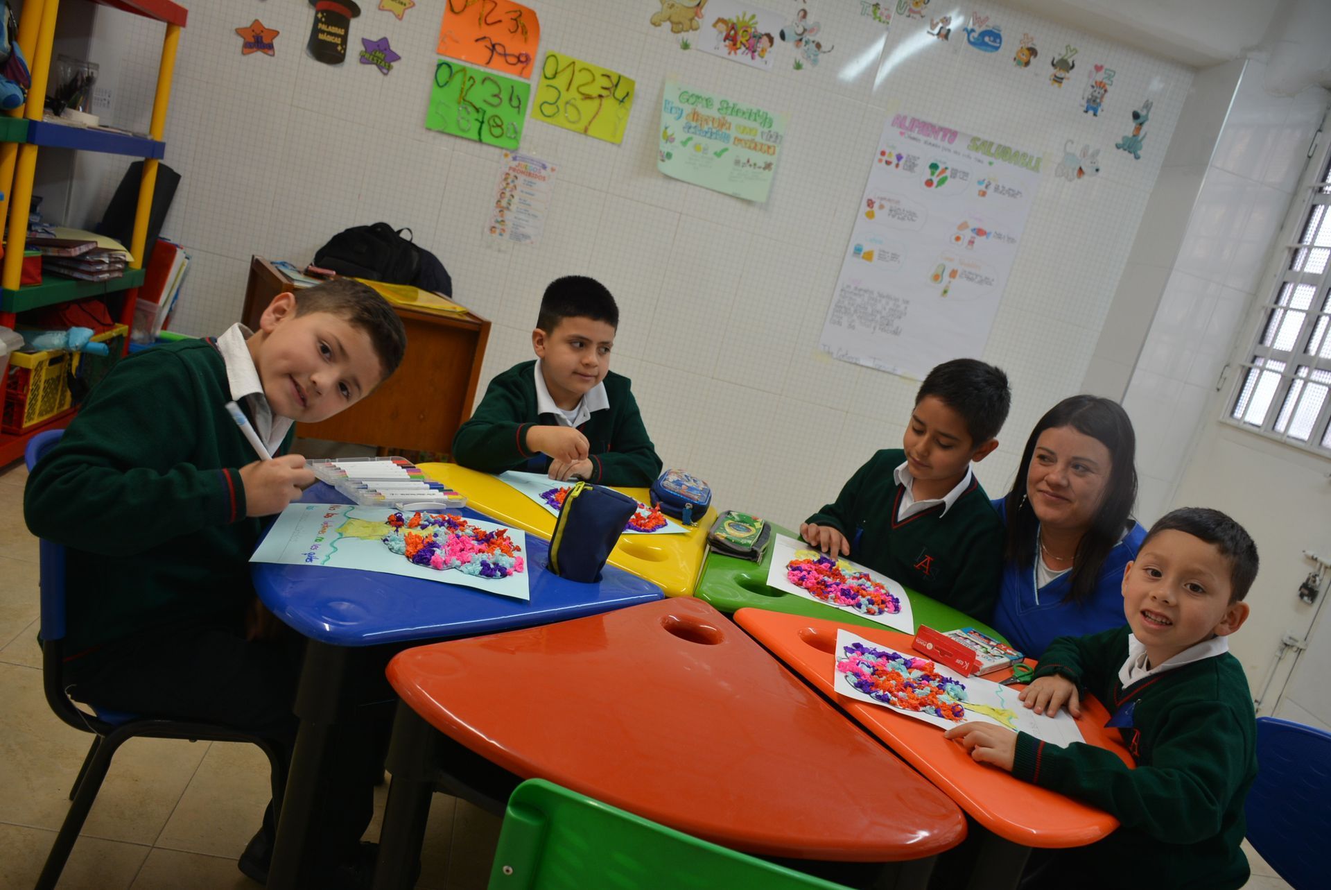 Niños sentados en mesas coloridas en un aula, trabajando en proyectos de arte y sonriendo a la cámara.