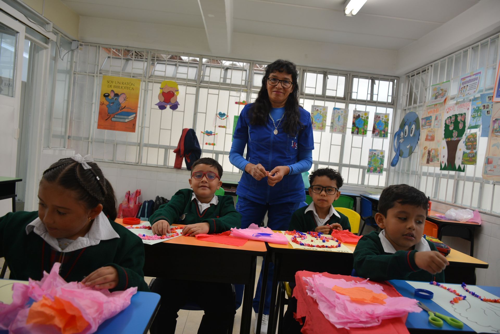 Alumnos y un profesor en un aula luminosa, con coloridas obras de arte en las paredes y manualidades de papel en los pupitres.