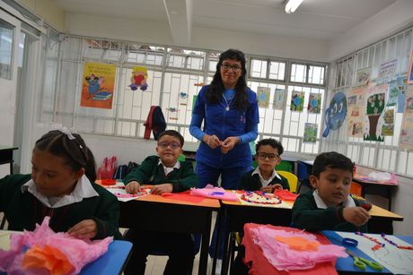 Alumnos y un profesor en un aula luminosa, con coloridas obras de arte en las paredes y manualidades de papel en los pupitres.