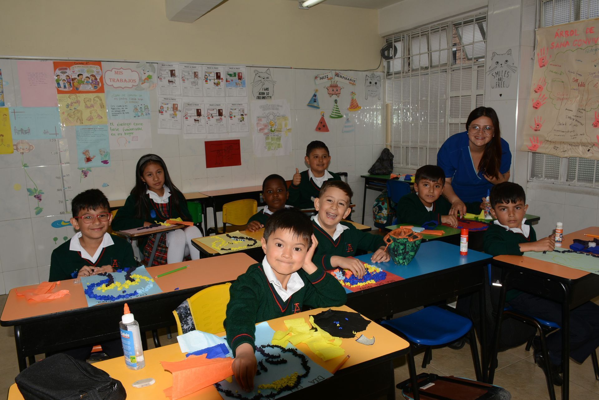 Alumnos sentados en sus pupitres en un aula, sonriendo junto a un profesor que se encuentra al fondo.
