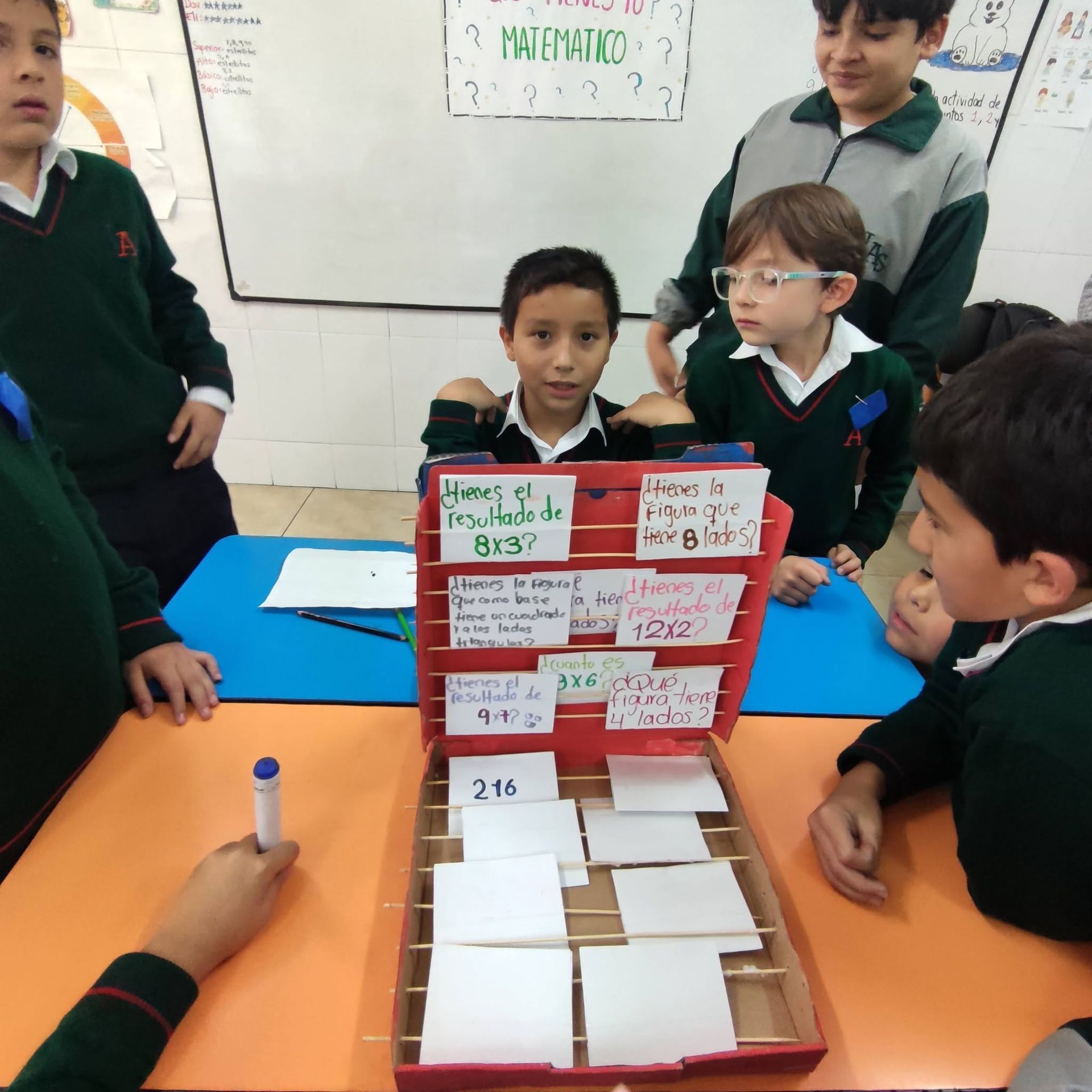 En un aula, niños con uniforme escolar se reúnen alrededor de un juego de mesa con cartas y bloques.