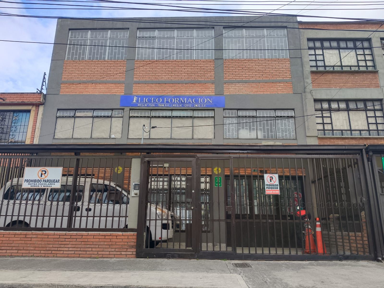 Vista desde la calle de un edificio de ladrillo de tres plantas con un letrero azul y una entrada con verja, bajo un cielo nublado.