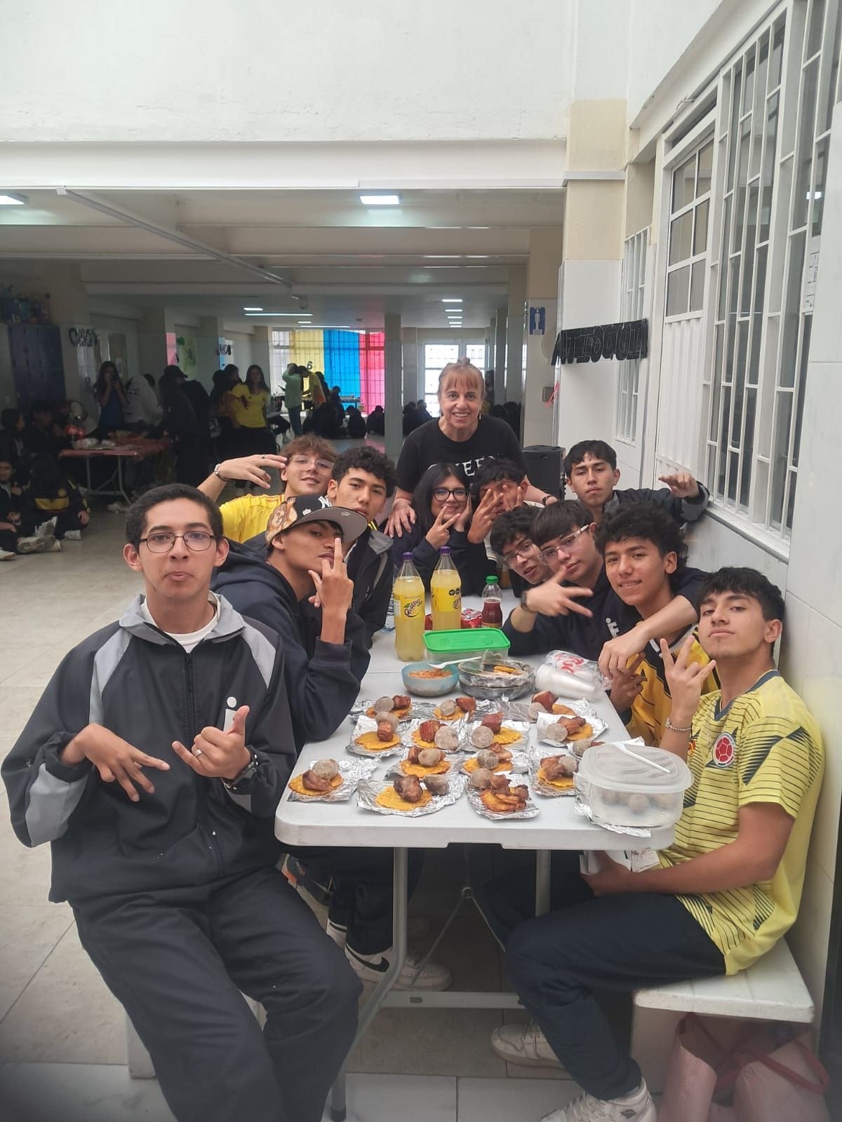 Un grupo de estudiantes se reunió alrededor de una larga mesa con comida en un luminoso pasillo interior, posando para una foto.