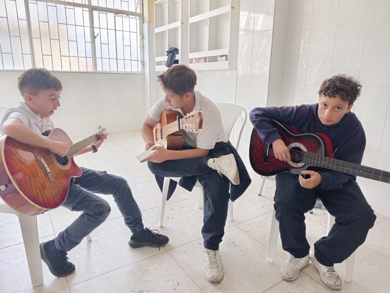 Tres personas sentadas en el interior, tocando guitarras acústicas en una habitación blanca y luminosa.