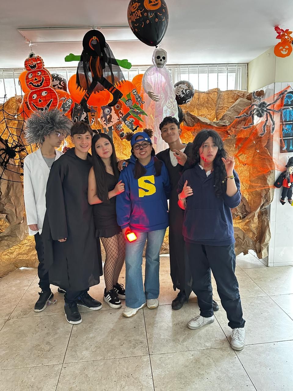 Grupo de seis personas posando en interiores con atuendos negros, con decoraciones de Halloween, hojas naranjas y globos de calabaza detrás.